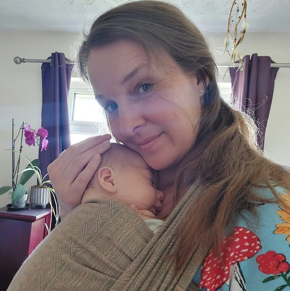 A woman with long brown hair holding a sleeping baby close to her face inside a room with a window, purple curtains, and an orchid plant on a wooden cabinet.