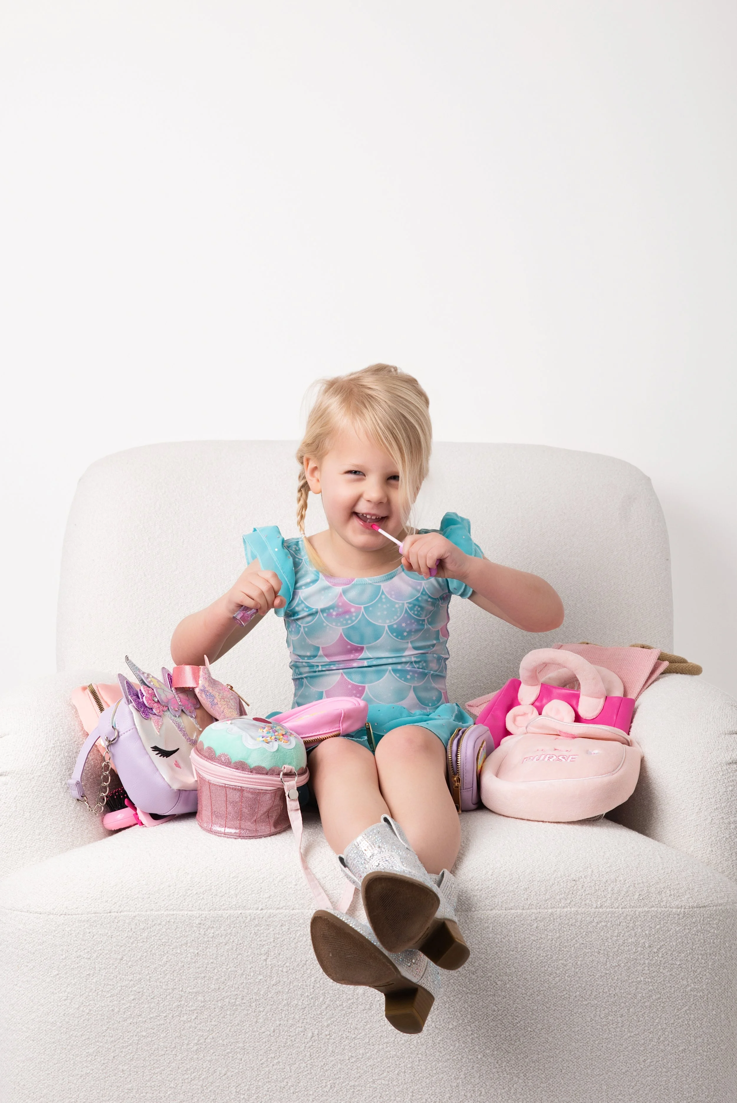 Young girl sitting on a white sofa with pastel-colored bags and toys, smiling and licking a lollipop.