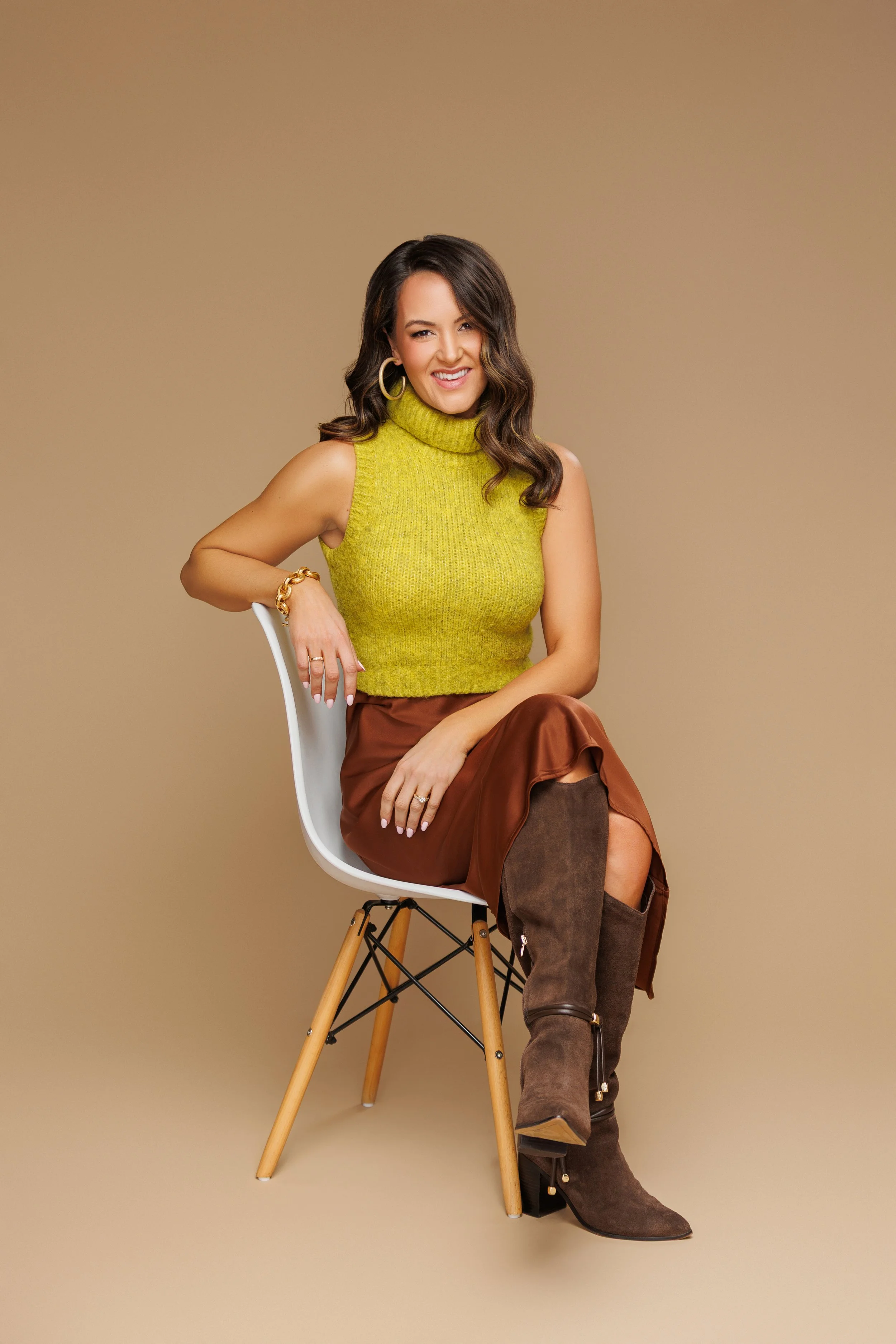 A woman sitting on a modern white chair with wooden legs, wearing a green sleeveless turtleneck sweater, a brown leather skirt, and brown knee-high boots. She is smiling and has wavy dark hair, large hoop earrings, and gold jewelry on her left wrist.