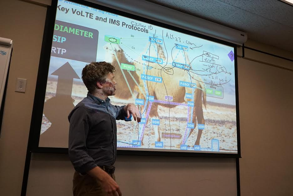 Owner of IRIS7, RZFeeser standing in front of a presentation slide at a conference or classroom. The slide displays a diagram of a camel with annotations and technical protocols related to 4G architecture, VoLTE, SIP, and IMS protocols.