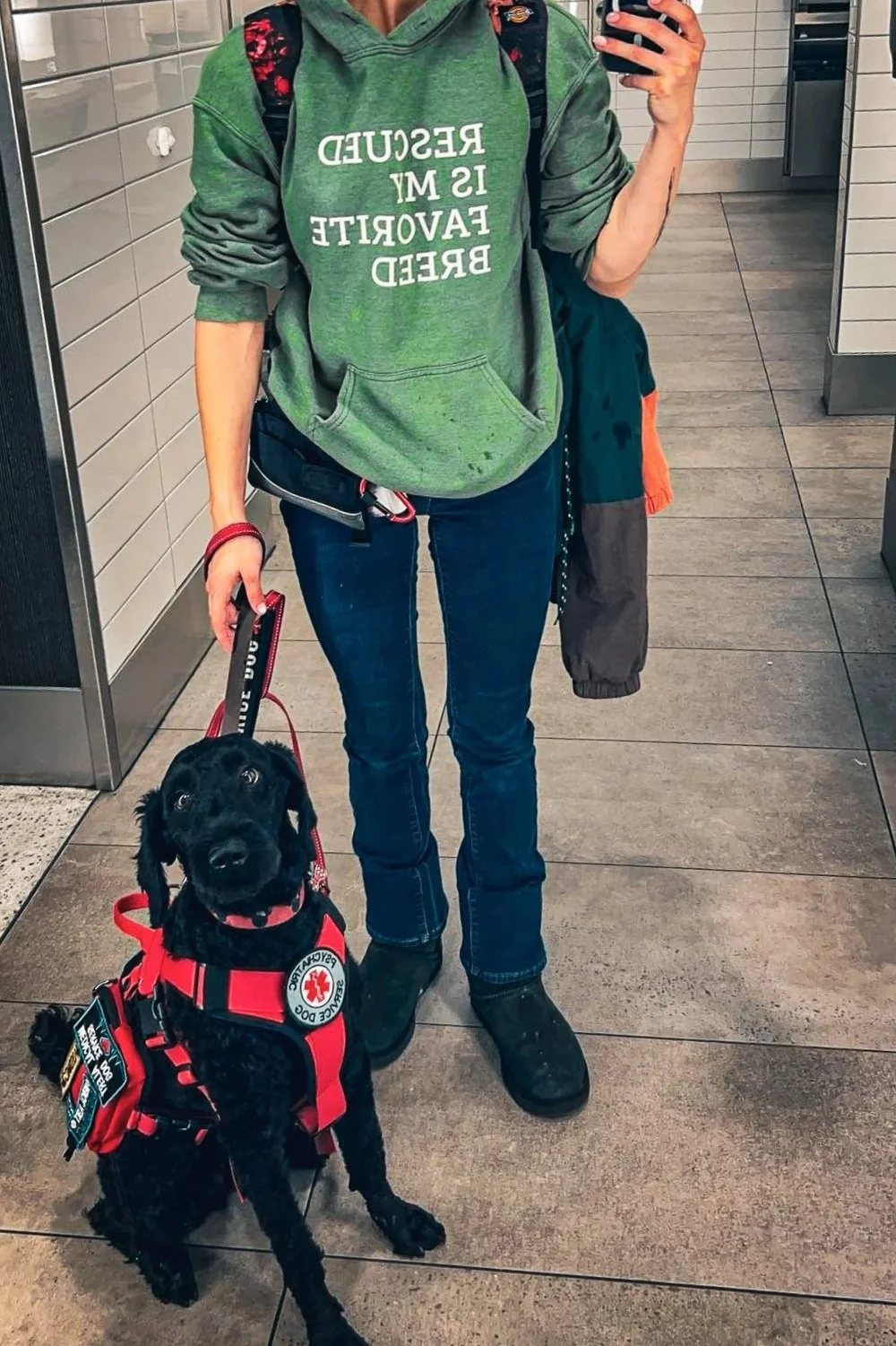 A person in a green hoodie with the words "RESCUED IS MY FAVORITE BREED" standing in a restroom with a black service dog in a red vest. The person is taking a mirror selfie, holding a phone with a black case in one hand and the dog’s leash with the other.