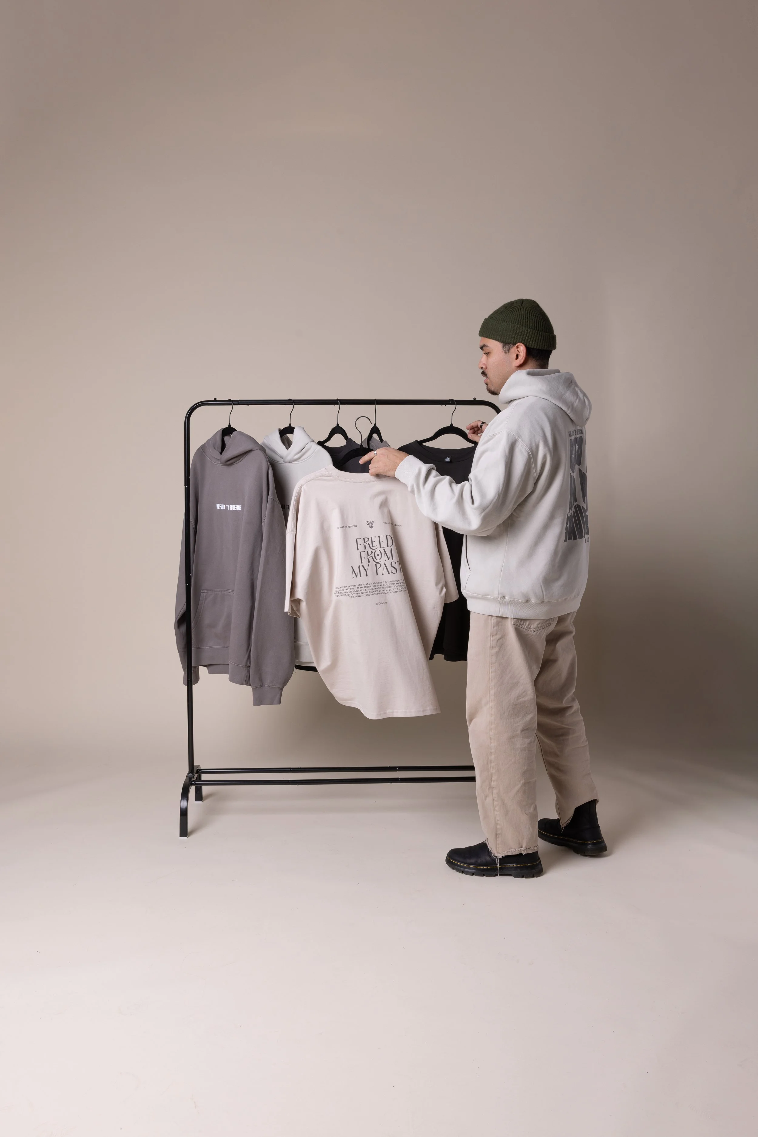 A man wearing a green beanie, white hoodie, beige pants, and black shoes looks at a clothing rack with hoodies and t-shirts in a neutral setting.