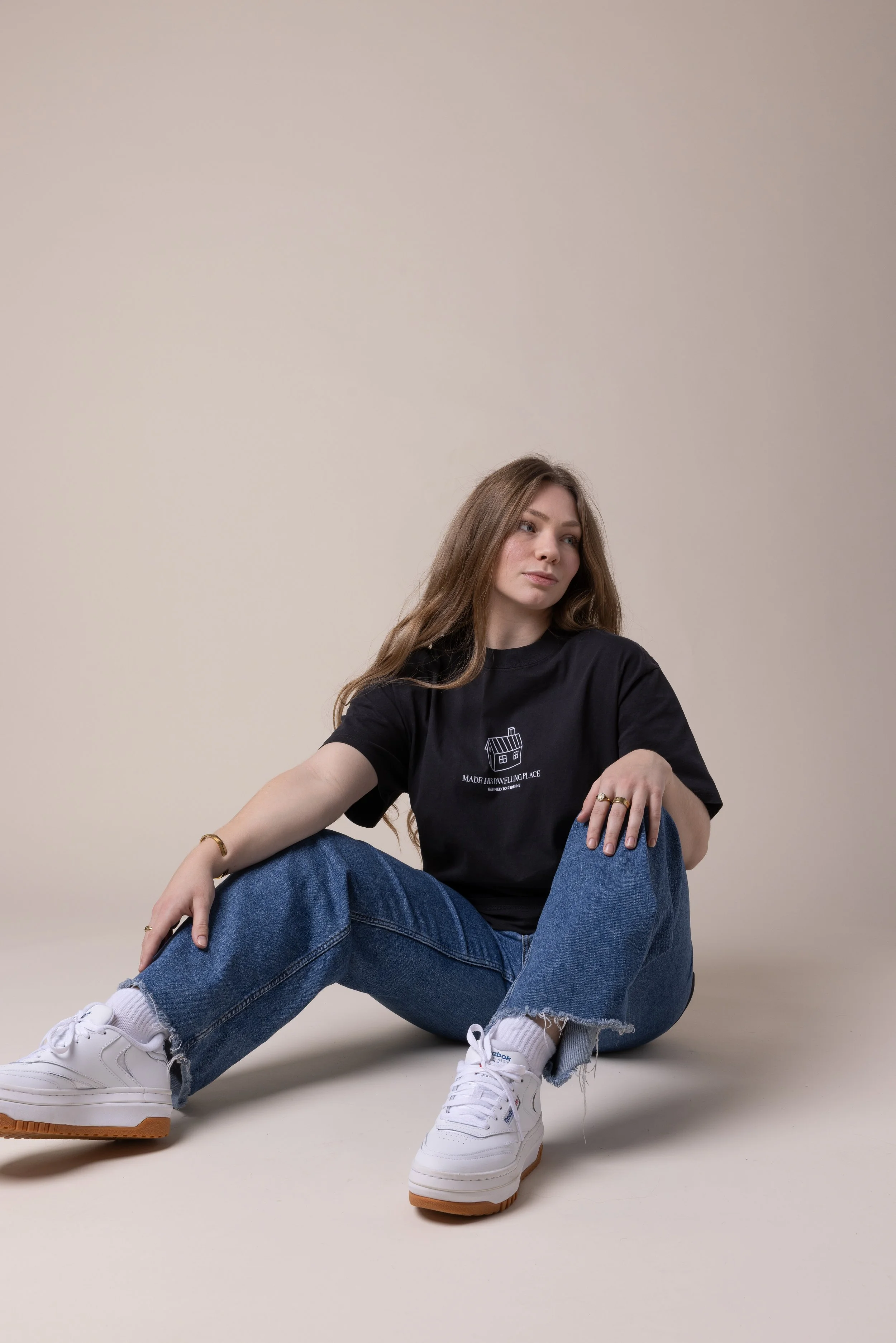 Young woman sitting on the floor against a plain background, wearing a black t-shirt, blue jeans, and white sneakers.