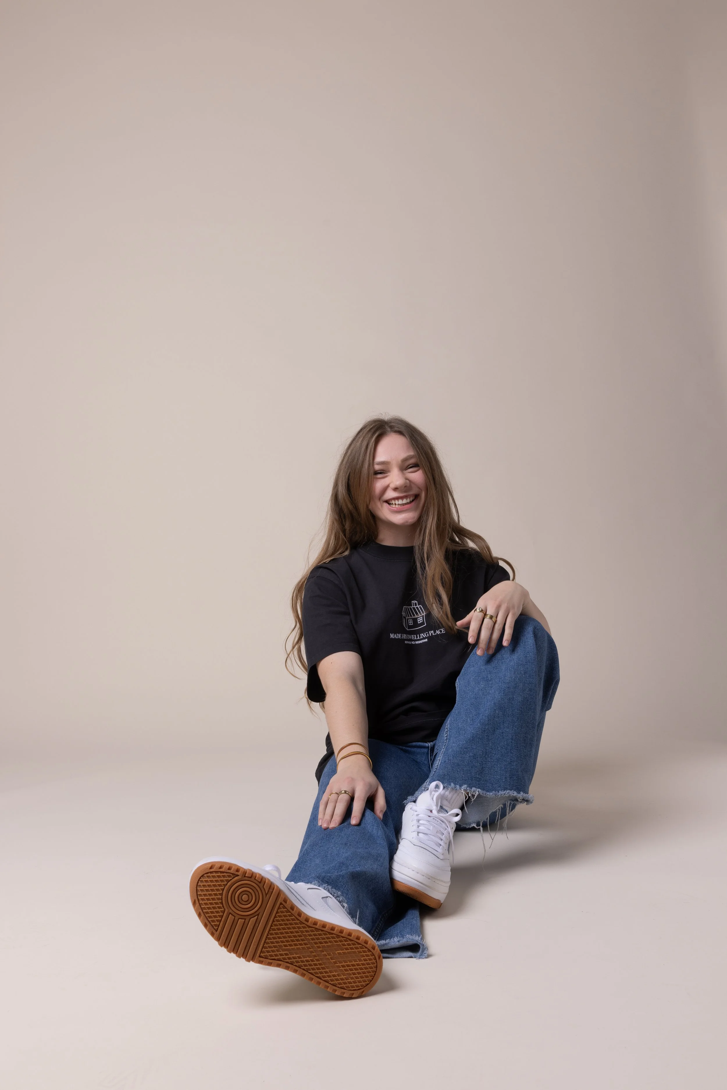 A young woman with long brown hair sitting on the floor, smiling, wearing a black t-shirt, blue jeans, and white sneakers.