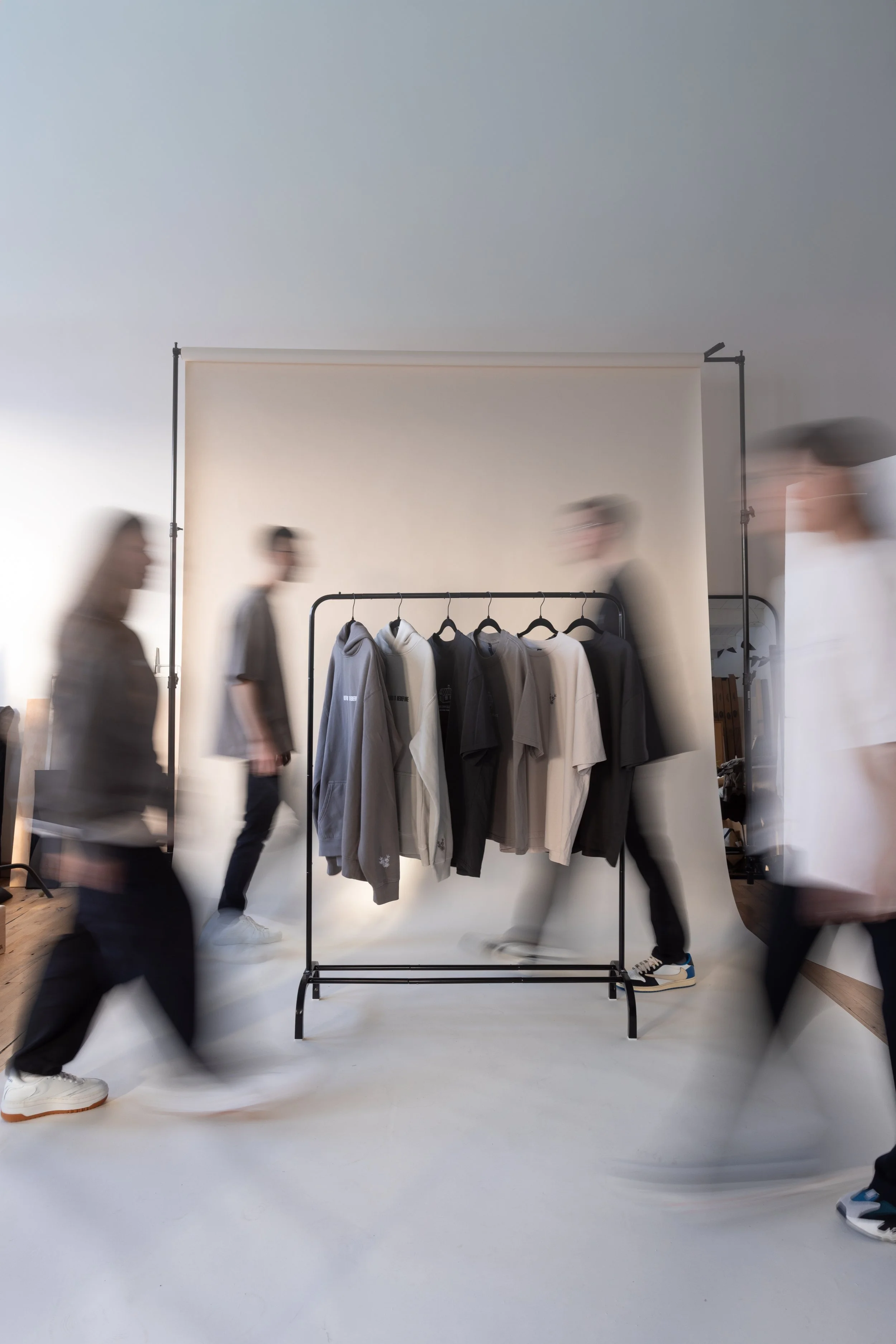 Clothing rack with gray, black, and beige T-shirts and hoodies in a room with blurred people walking by.