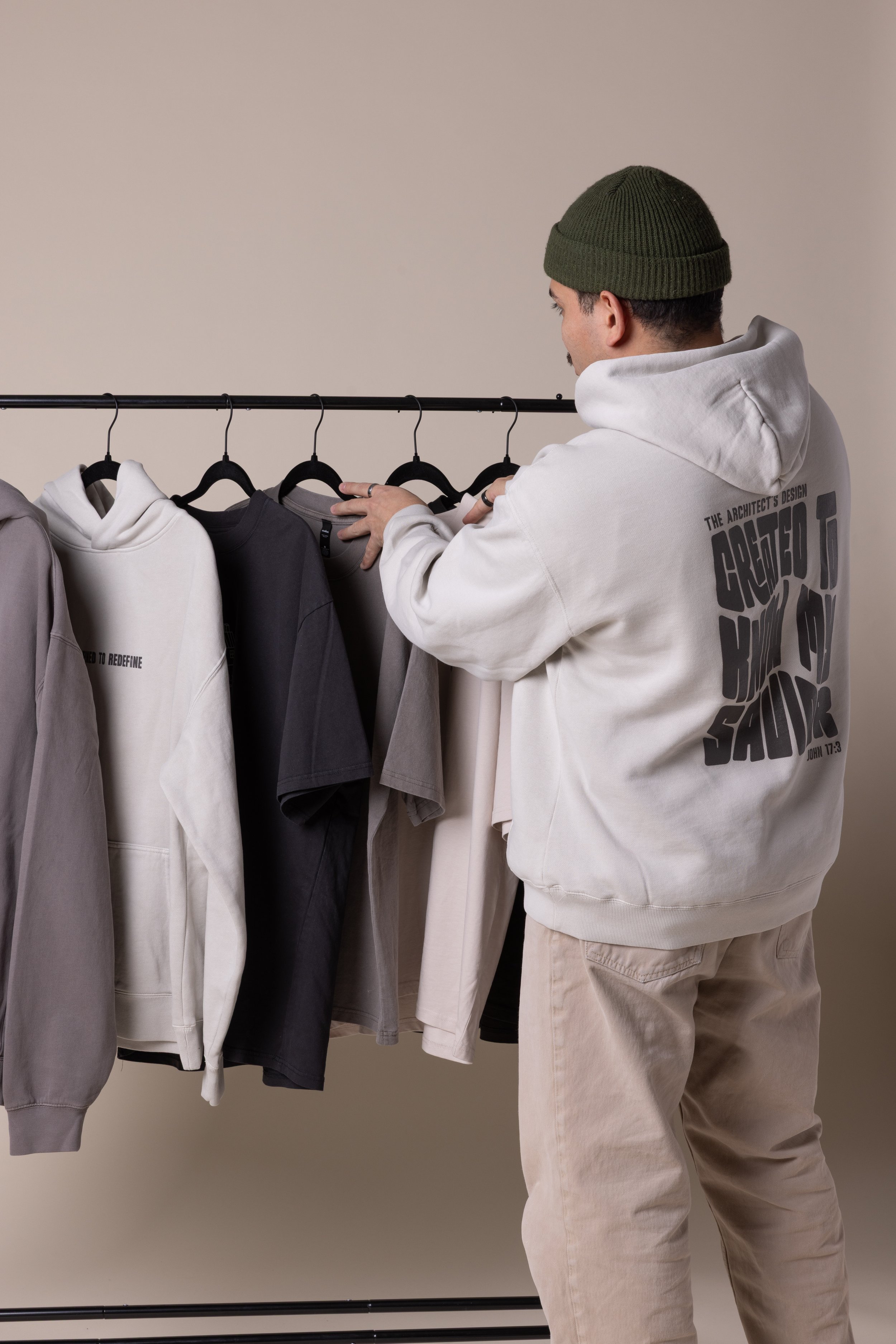 A man wearing a green knit cap, white hoodie, and beige pants browsing a clothing rack with various colored hoodies and t-shirts.
