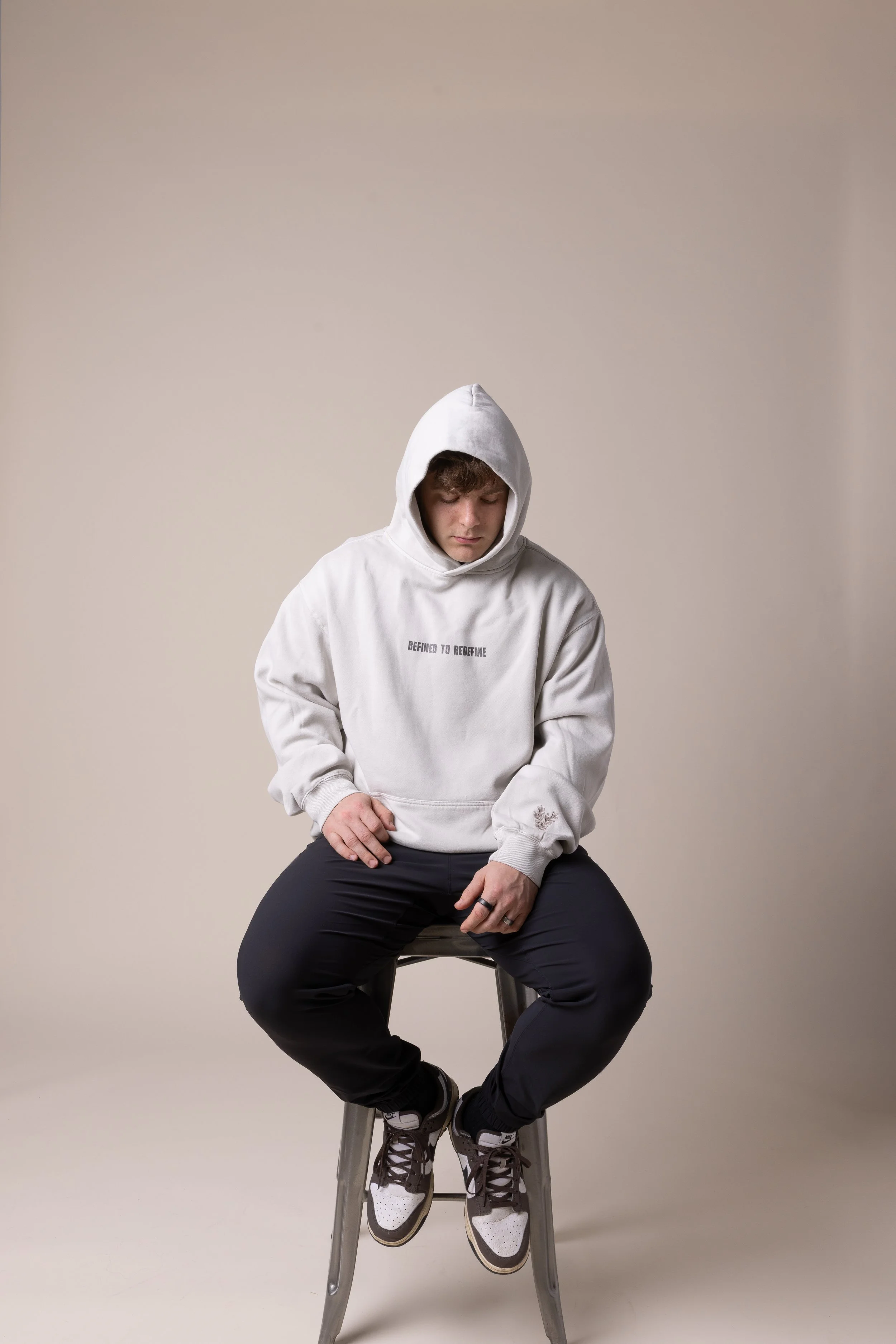 A young man wearing a white hoodie with the text 'Refined to Redefine' sits on a stool against a plain beige background, looking down with a serious expression.