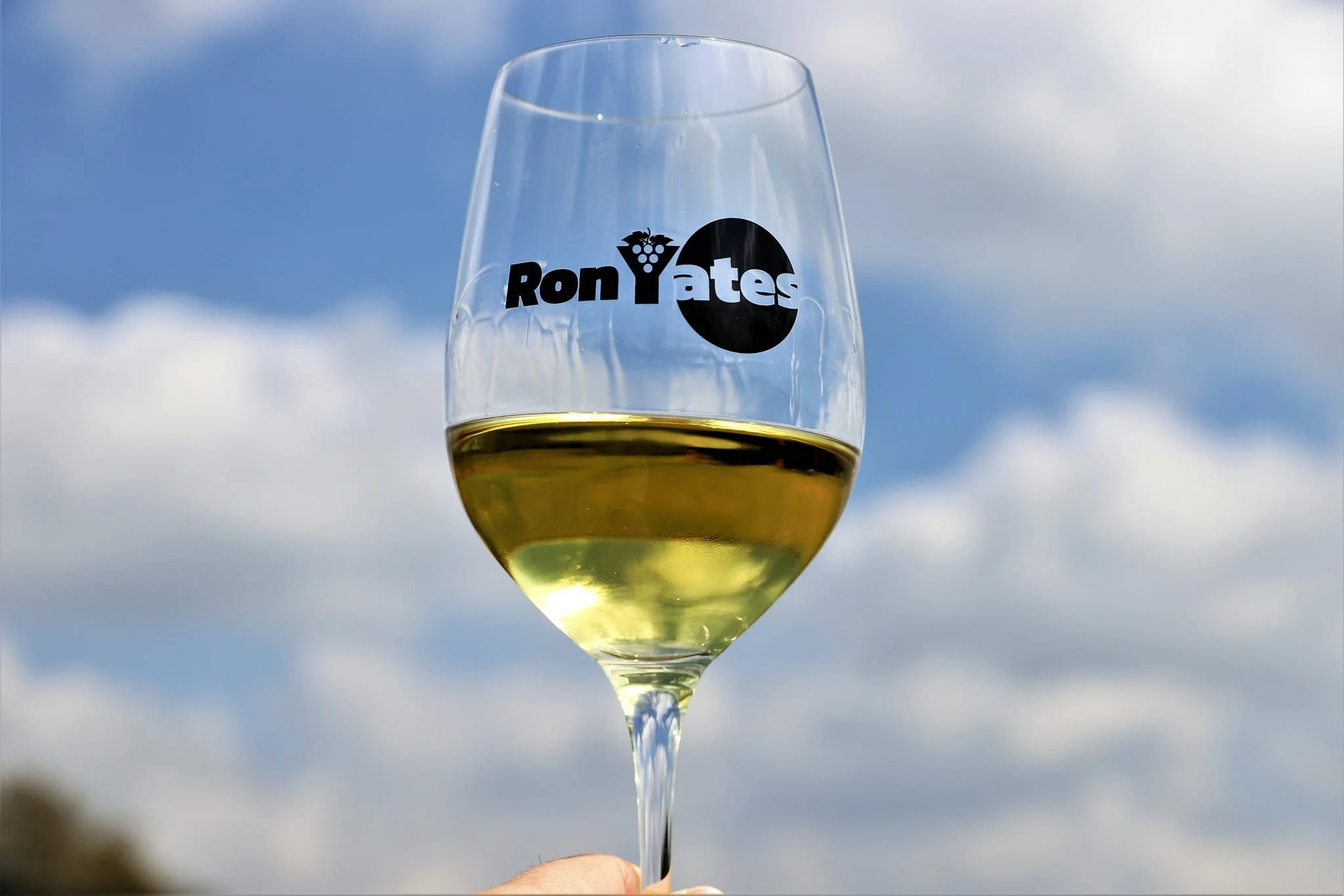 A glass of white wine held up outdoors with a cloudy sky in the background. The glass has a logo that reads 'Ron Yates' with a grapevine icon and a black circle.