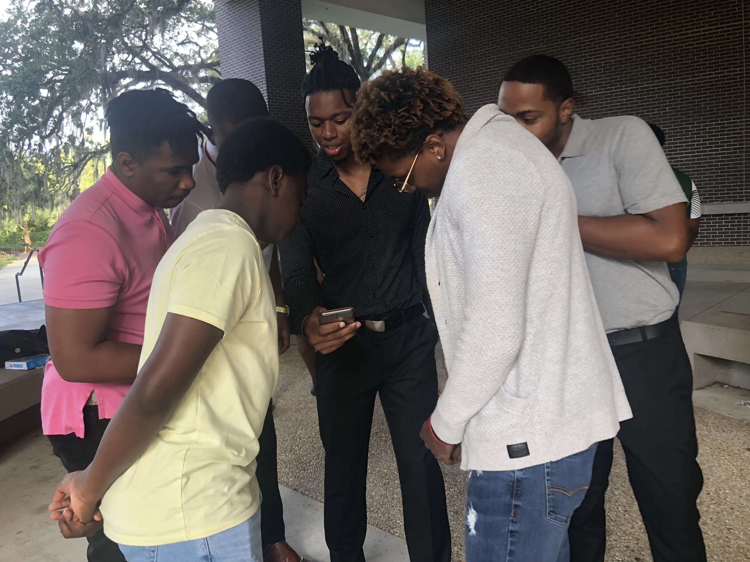 Group of six young people, three men and three women, standing closely together outdoors, looking at a phone held by one woman, in front of a building with brick walls and trees in the background.