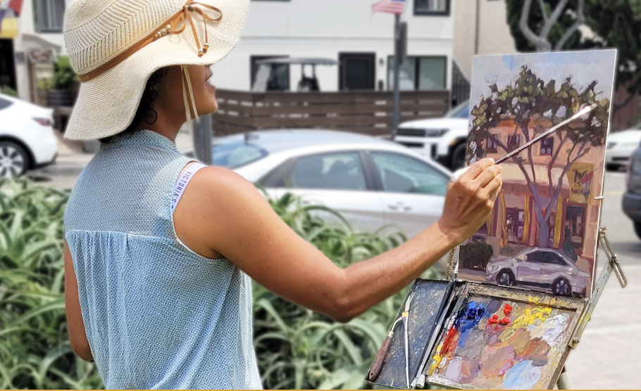 Person painting outdoors, wearing a striped sweater, holding a paintbrush and palette on an easel, with blurred greenery in the background.