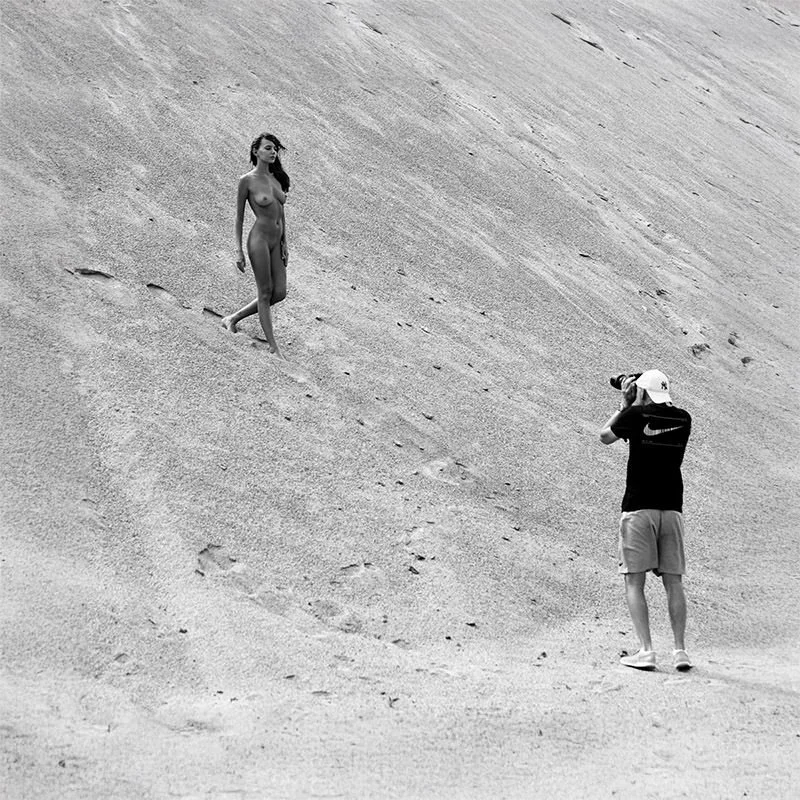 A woman without clothes or a shirt and a man in a cap and shorts taking a photo of her on a sandy incline. The woman stands on the sand, and the man is standing and taking photos with a camera.