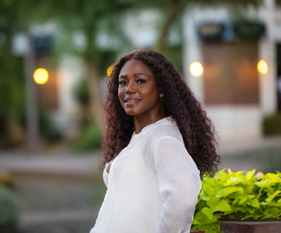 Portrait of a woman with long curly hair wearing a white shirt, standing outdoors with a blurred background of trees, lights, and a building.