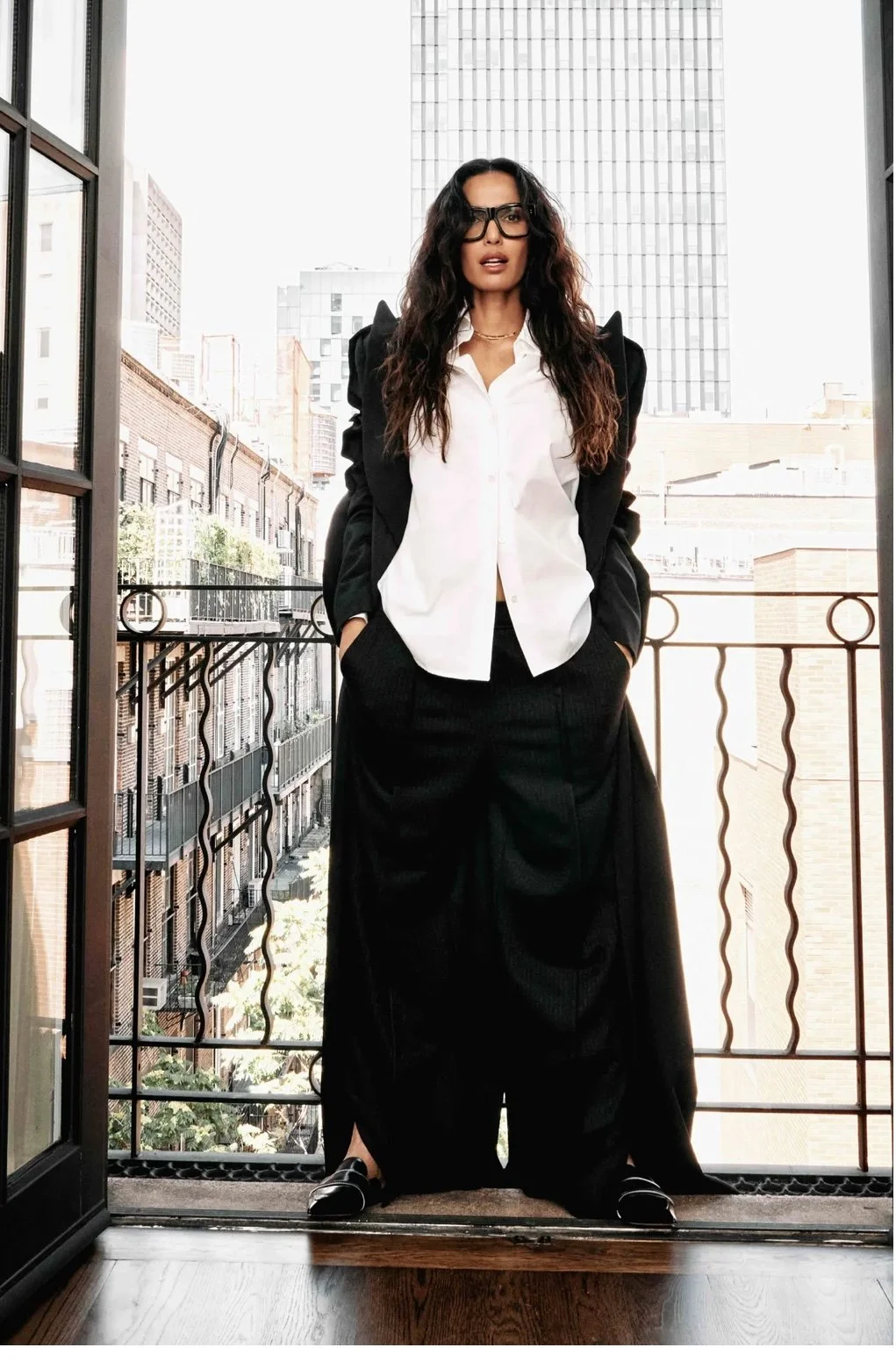 padma lakshmi for Marie claire standing against an open balcony styled in a white button down, and a deconstructed black suit with wide frame sunglasses