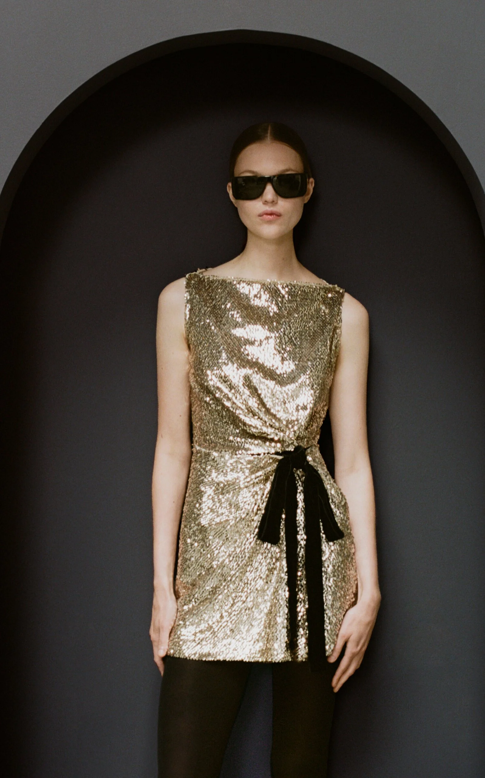 moda operandi holiday 2025 model wearing a short gold sequined mini dress cinched with a black velvet bow, styled with semi opaque black tights and dark black rectangle-framed sunglasses