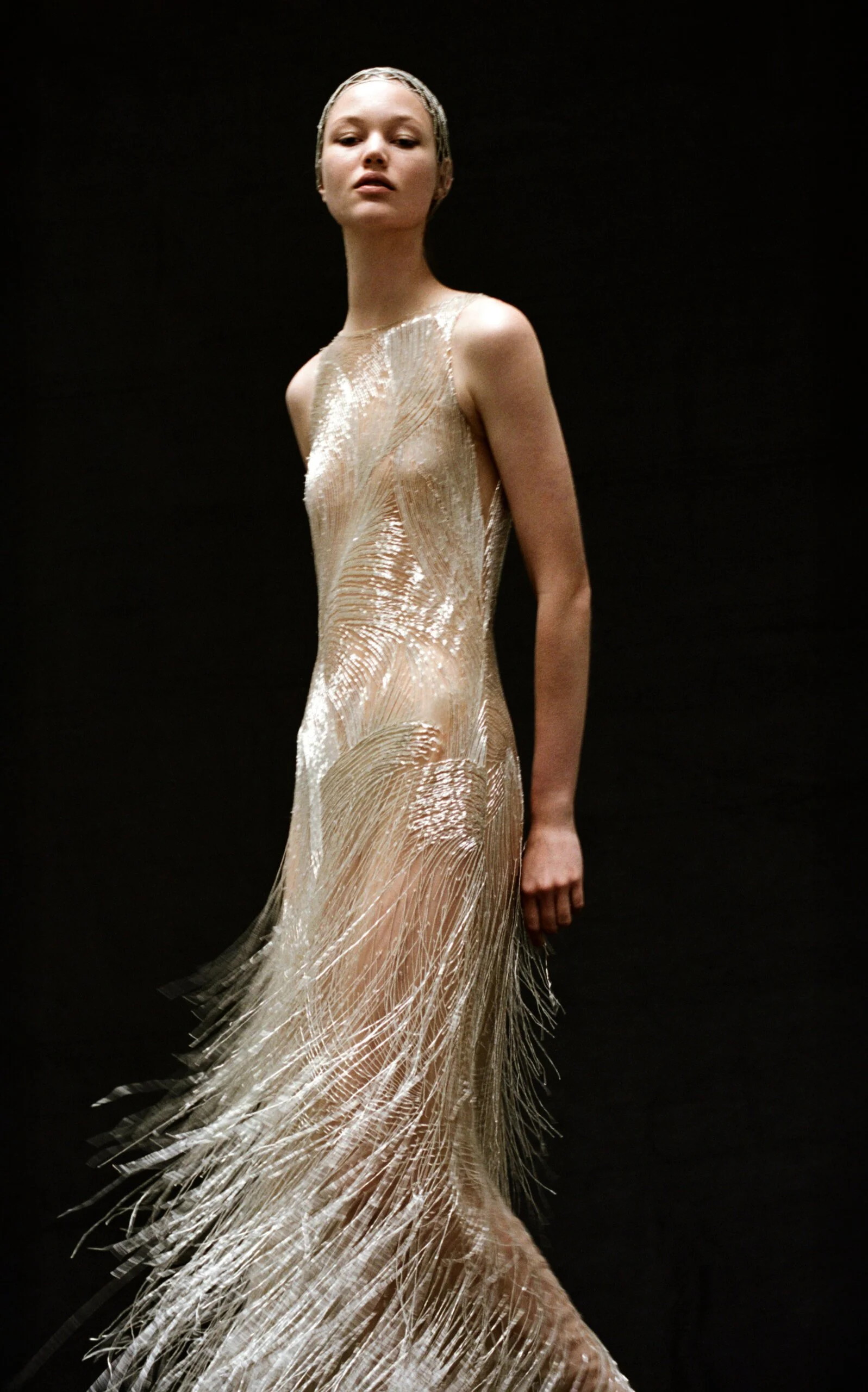 moda operandi holiday 2025, model wearing a sheer gold flapper dress with beading and feathers against a black background