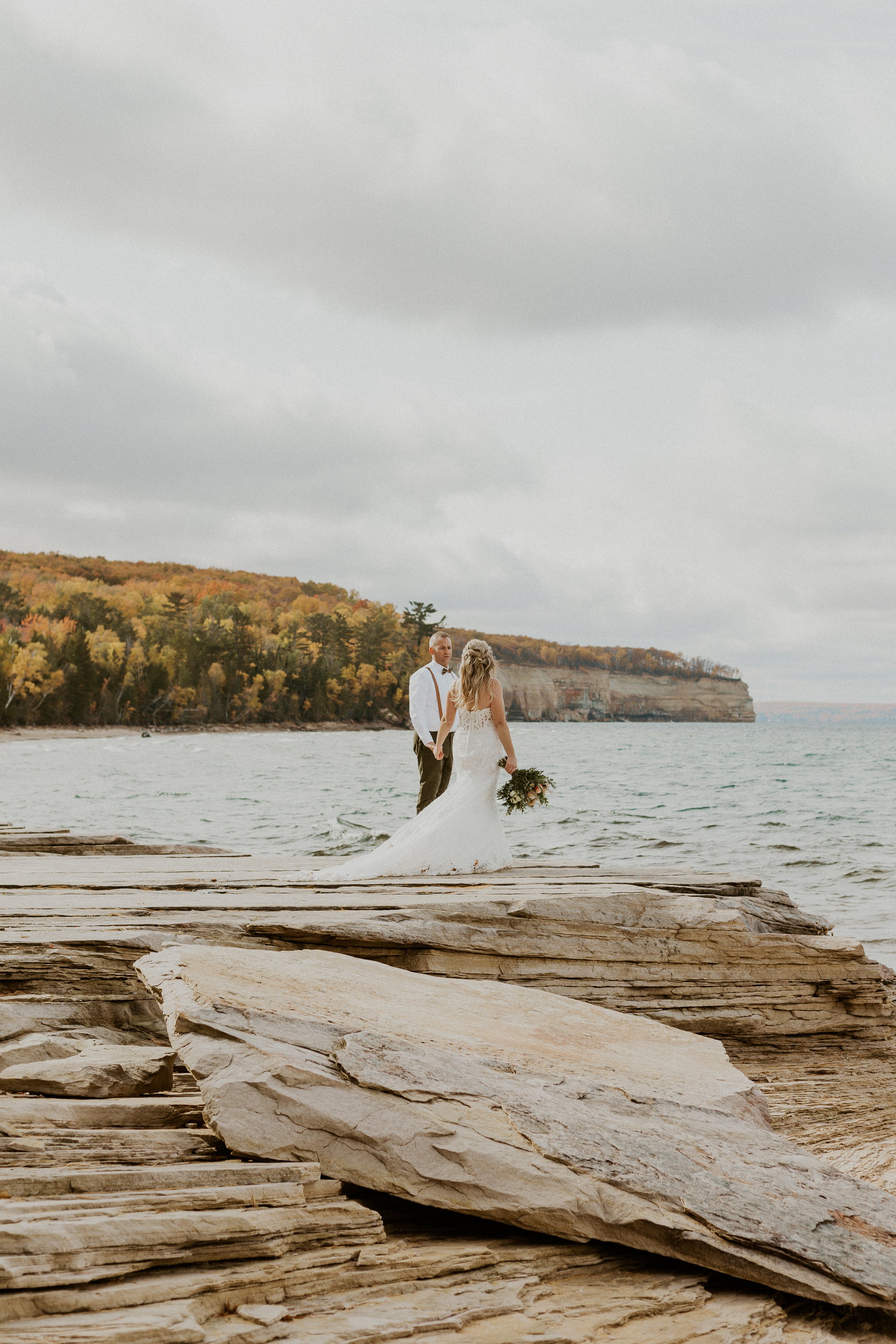 A bride and groom holding hands near the Pictured Rocks shoreline on a cloudy day, with rocky terrain and a forested cliff in the background.