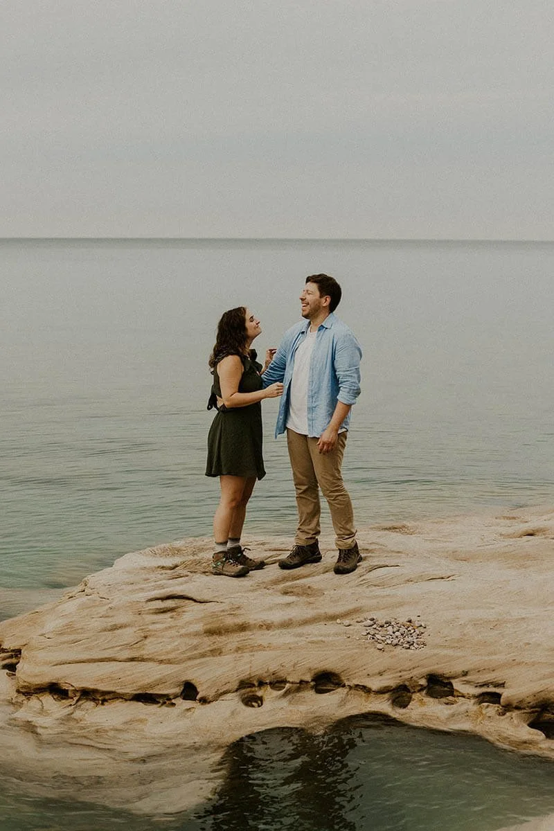 A couple standing on a large rock by a calm lake, smiling at each other.