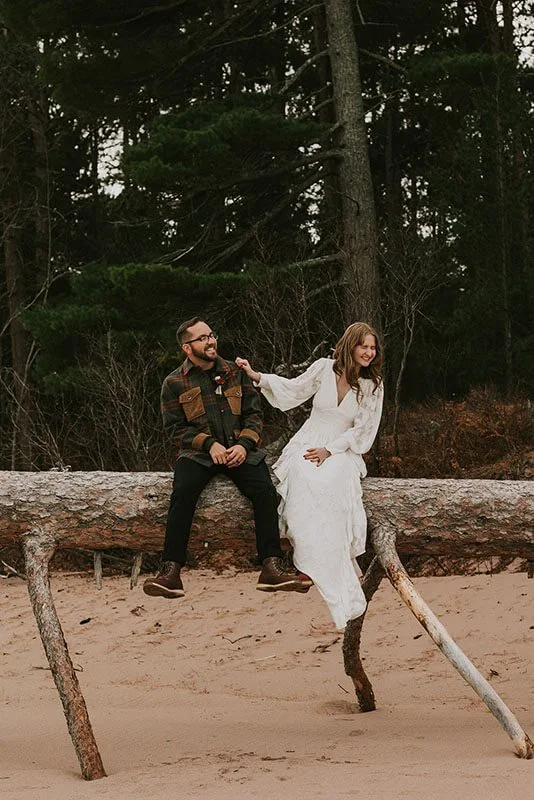 A man and woman sitting on a fallen tree trunk on a sandy beach, surrounded by tall trees, with the woman in a white dress and the man in dark pants and a plaid jacket, smiling and enjoying each other's company.
