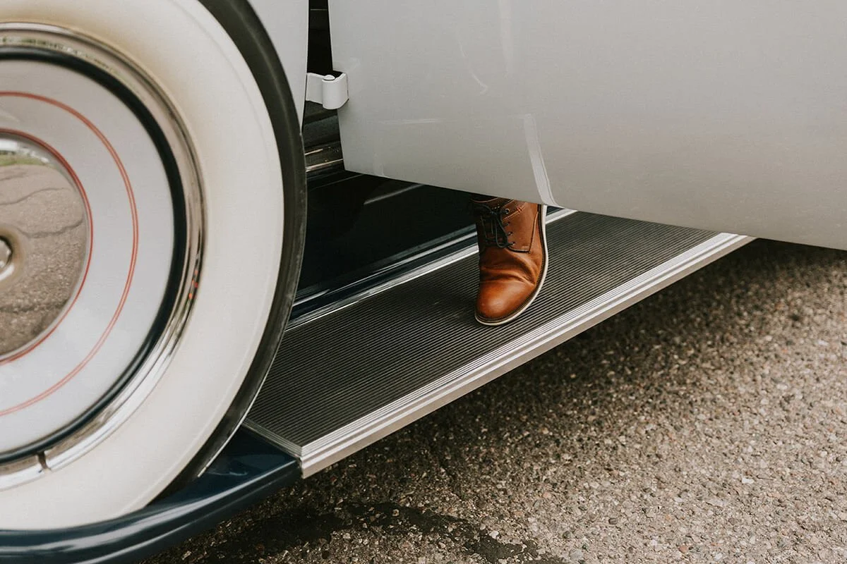 A person wearing brown leather boots stepping out of a vintage white car with large white-walled tires onto a paved surface.