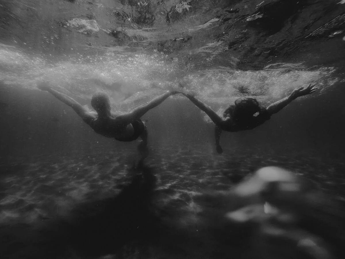 Two people holding hands and swimming underwater in a black and white photo, creating a heart shape with their arms.