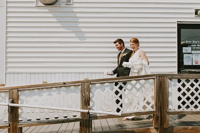Bride and groom walking together on a wooden outdoor deck, smiling, with a white building with siding and a window behind them.