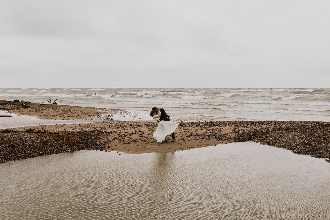 A couple in wedding attire sharing a romantic embrace and kiss on a cloudy beach in Michigan's Upper Peninsula with waves and driftwood in the background.