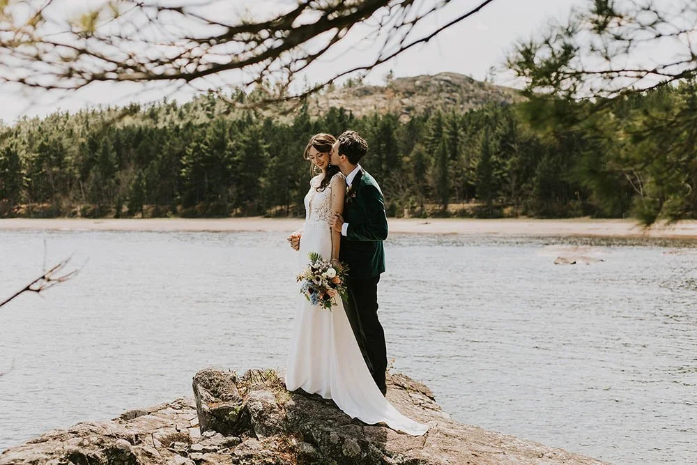 A bride and groom standing on a rock by a river, with trees and mountains in the background, sharing a moment of affection.