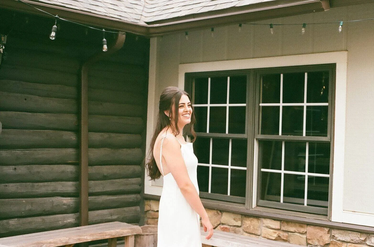 A woman in a white dress smiling and standing outside near a window with a wooden log cabin of Barrel and Beam Brewery wall behind her.