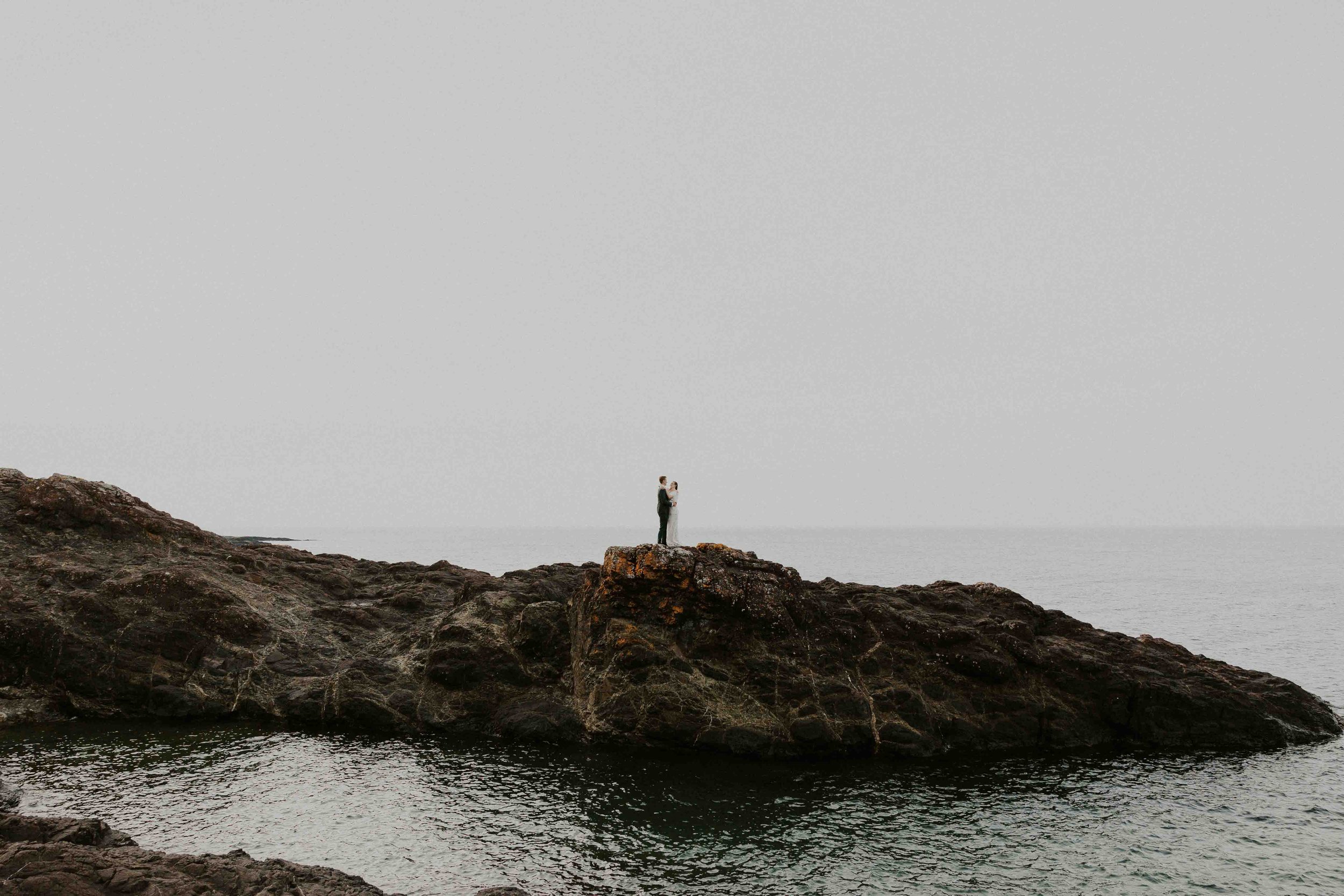 A couple in wedding attire stands on a rocky outcrop by Lake Superior on Black Rocks in Marquette, embracing each other with a foggy sky overhead.