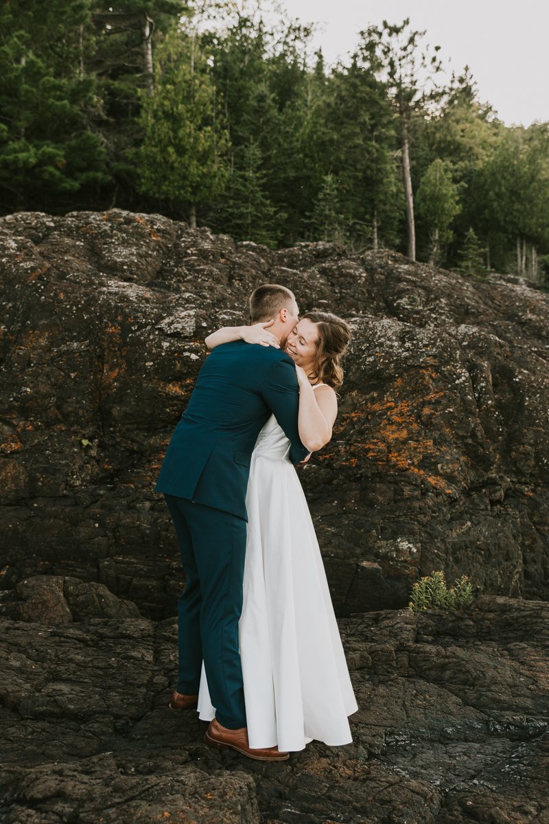 A couple dressed in formal attire sharing a kiss outdoors on a rocky terrain in Marquette Michigan with a forest in the background.