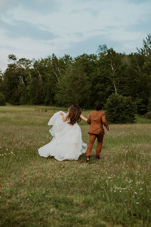 Bride and groom walking hand in hand through a grassy field with trees in the background.