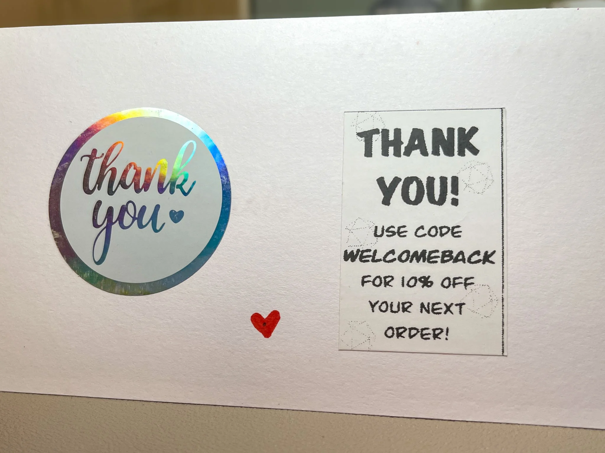 A photo of the back of an index card. On the left is a holographic thank you sticker, and on the right is a sticker with the text "Thank you! Use code WELCOMEBACK for 10% off your next order!"
