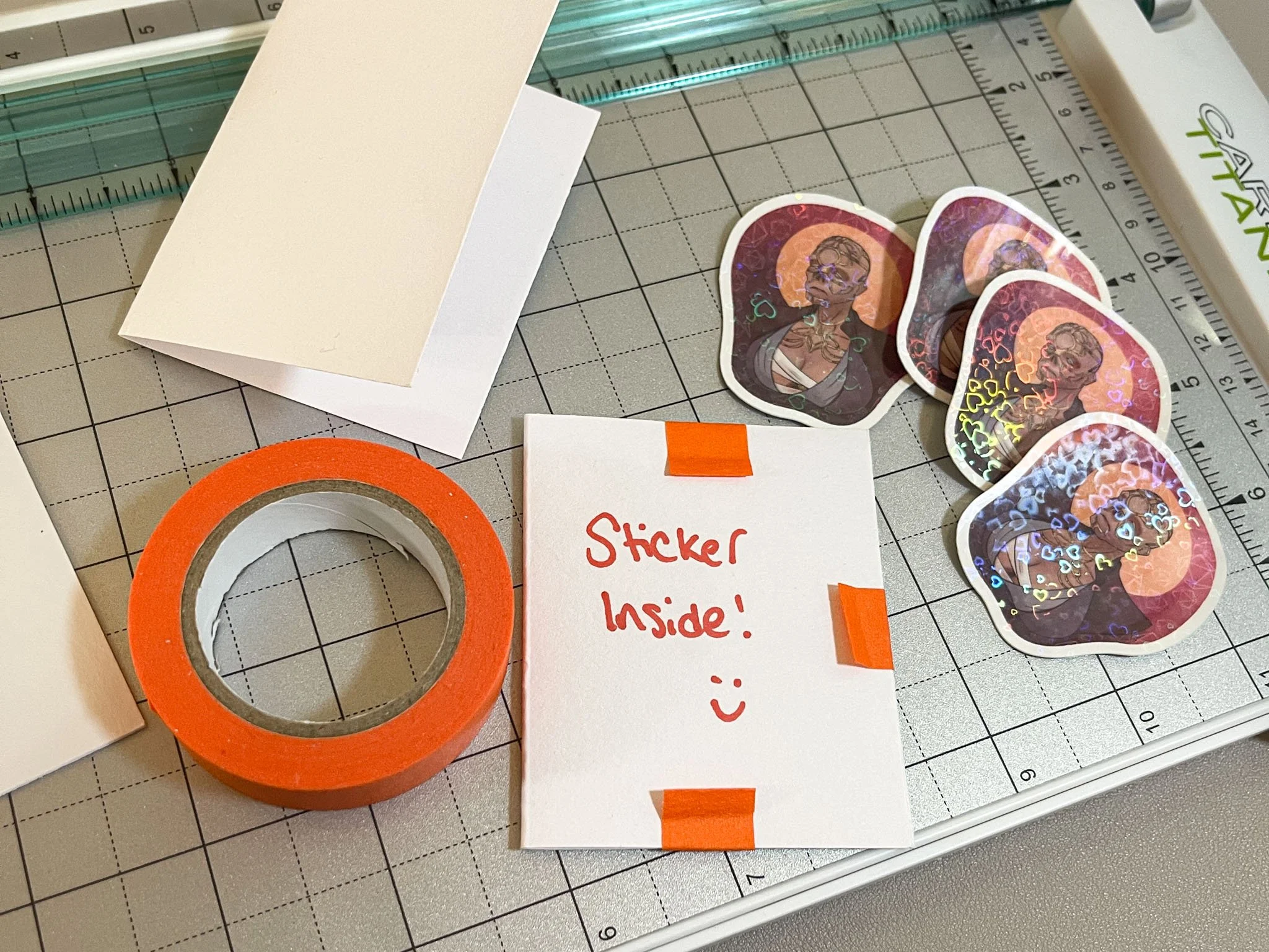 A photo of several stickers and stationery items on top of a ruled paper cutter. There is a folded index card, a roll of orange masking tape, a pouch with the text "Sticker inside! :)", and four holographic Withers Big Naturals stickers.