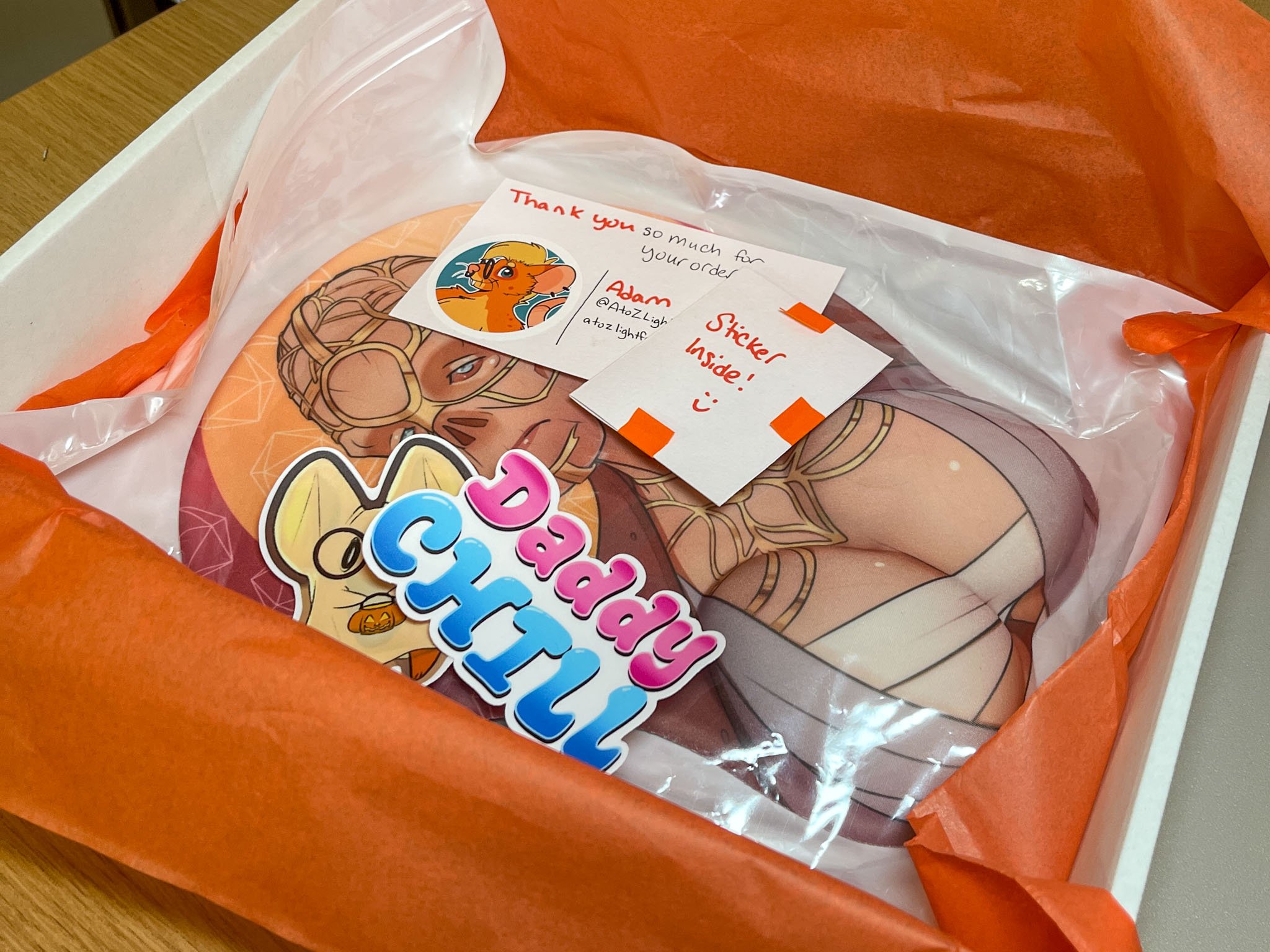 A photo of a packaged order, containing a Withers Big Naturals 3D mousepad, a thank you note, a sticker pouch, a rat ghost sticker, and a Daddy Chill sticker. Everything is in a white box on top of a sheet of orange tissue paper.