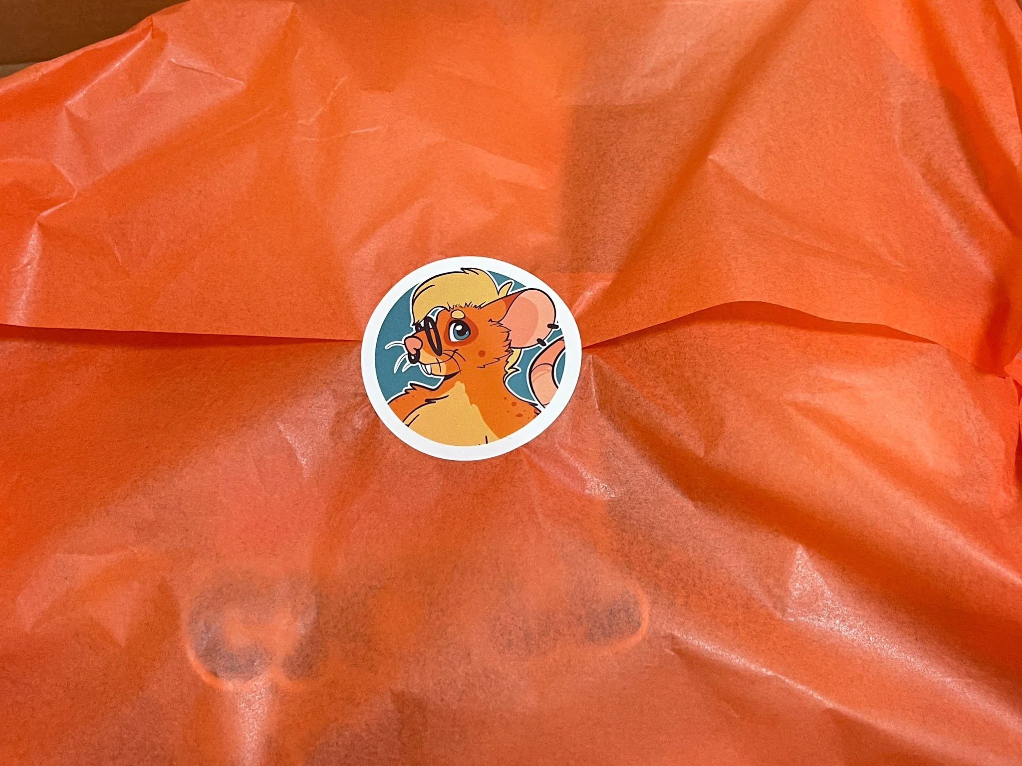 A photo of orange tissue paper sealed with a sticker of Clove, an orange rat character.