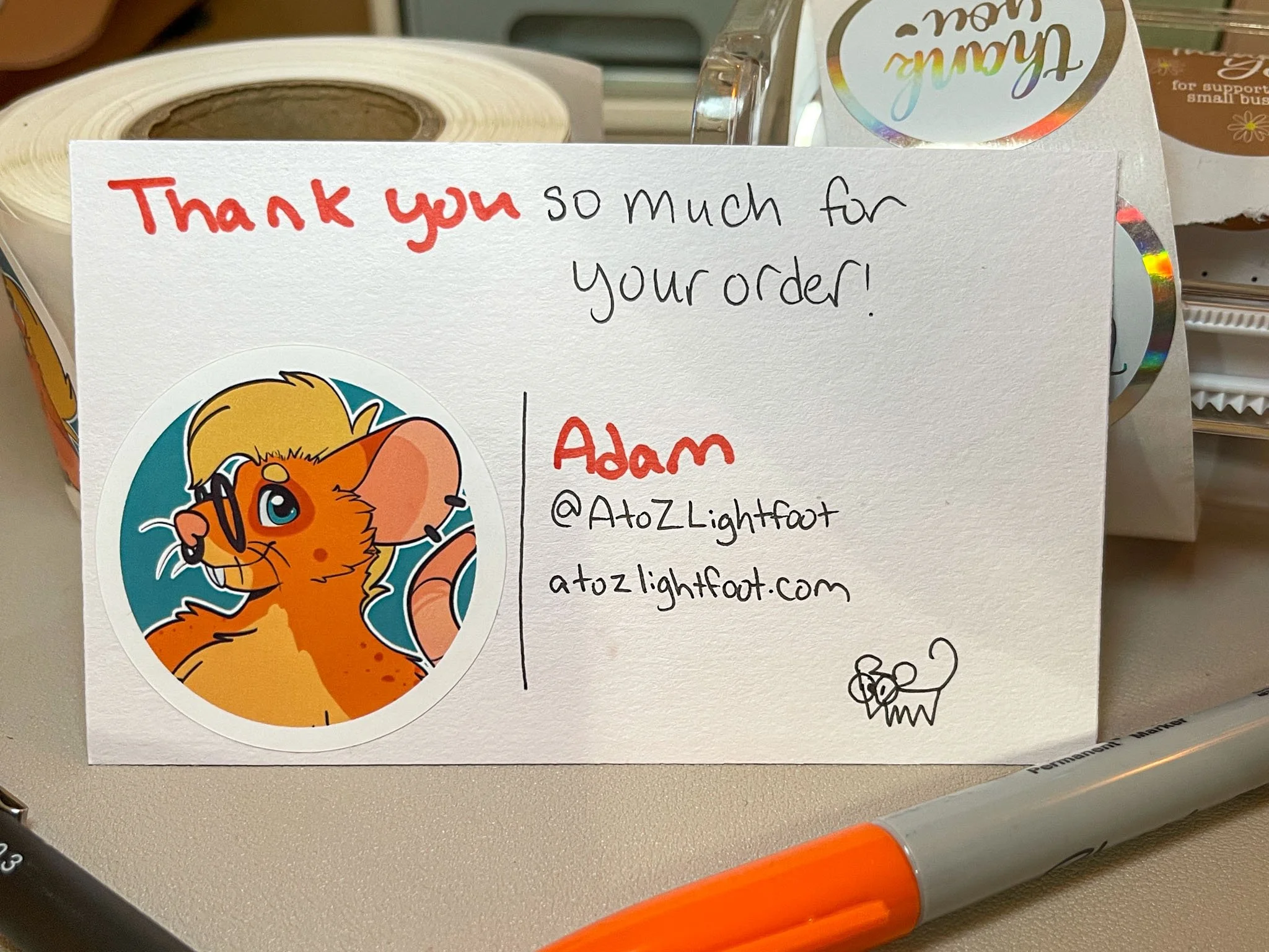A photo of an index card containing a circular sticker with Clove, an orange rat character, as well as the text "Thank you so much for your order! | Adam | @AtoZLightfoot | atozlightfoot.com". At the bottom of the card is a simple drawing of a rat.