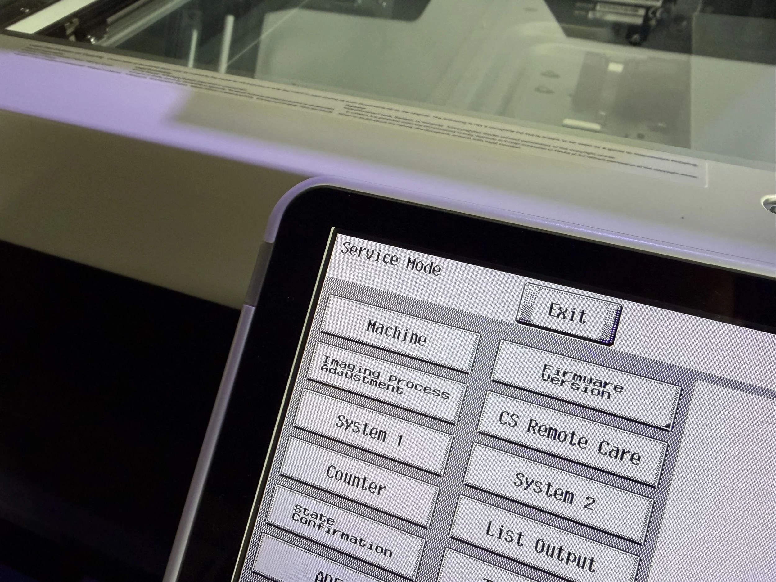 Close-up view of copier screen displaying a menu with options including 'Service Mode,' 'Exit,' and various system settings, with a background that shows a white electronic device with a glass surface and components inside.