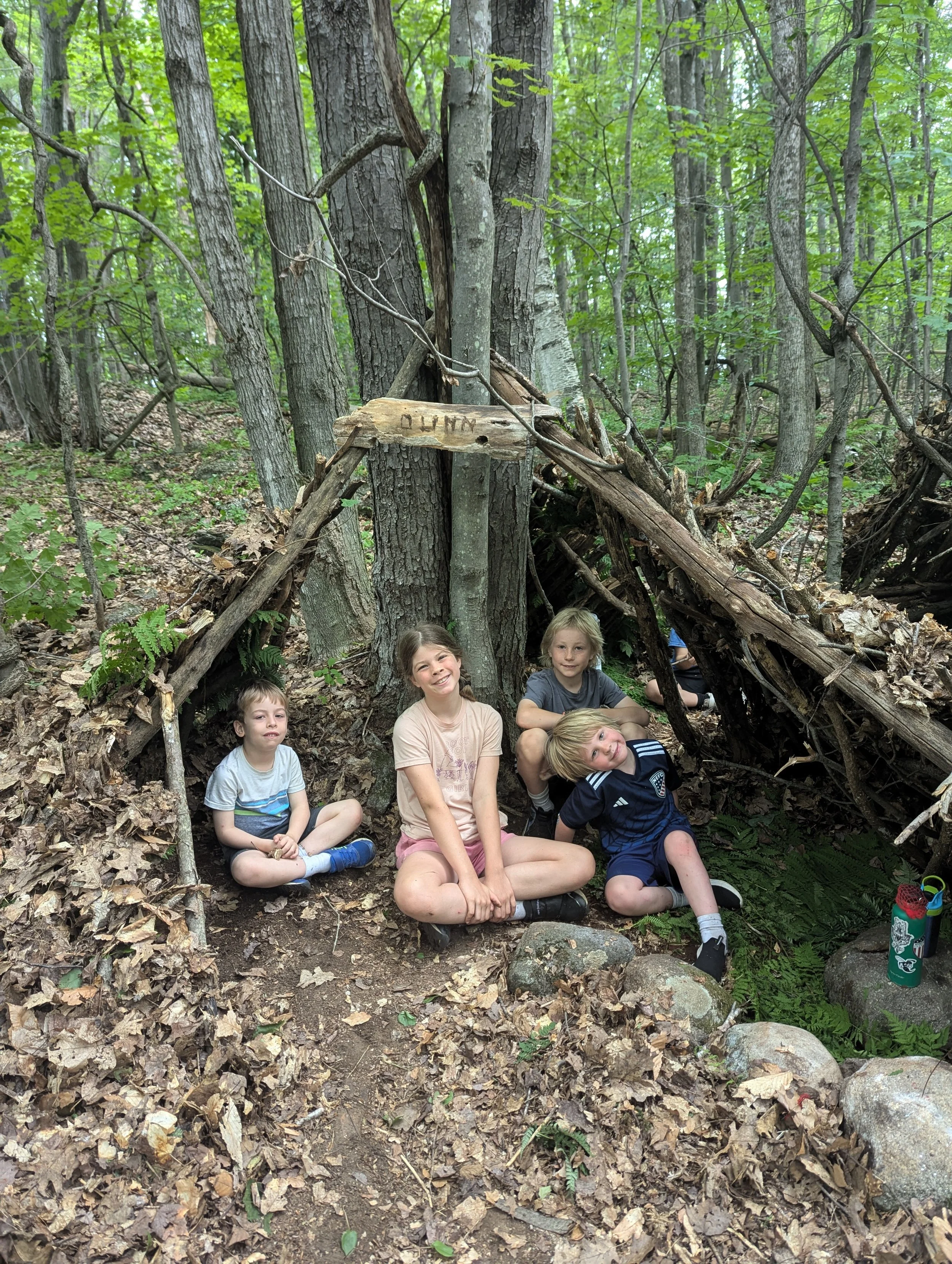 Four children sitting on the forest ground in front of a makeshift shelter built from sticks and leaves, with trees and green foliage in the background.
