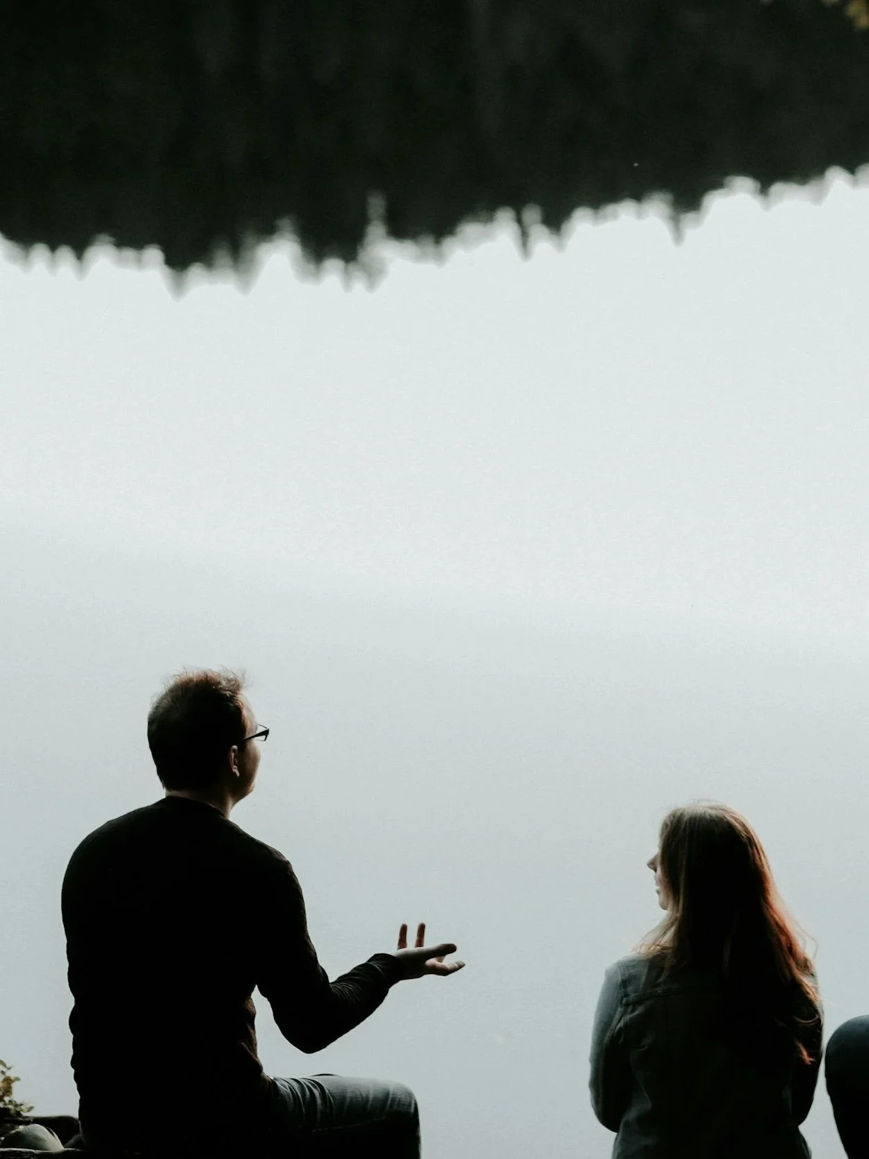 A man and a woman sitting outdoors near a body of water, facing each other, with the man gesturing as he speaks, the scene is calm and reflective, possibly on a cloudy day.