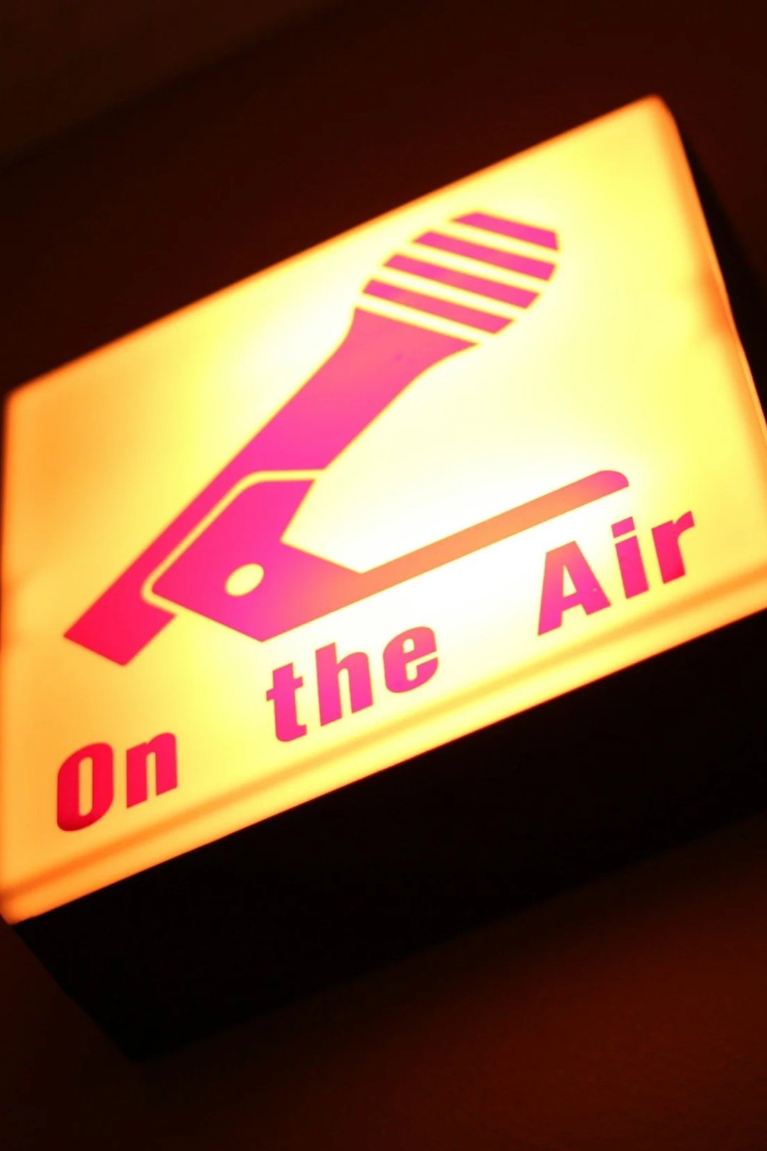 Illuminated sign with a pink and purple design of a hand turning a knob, along with the text "on the Air" in pink and purple.