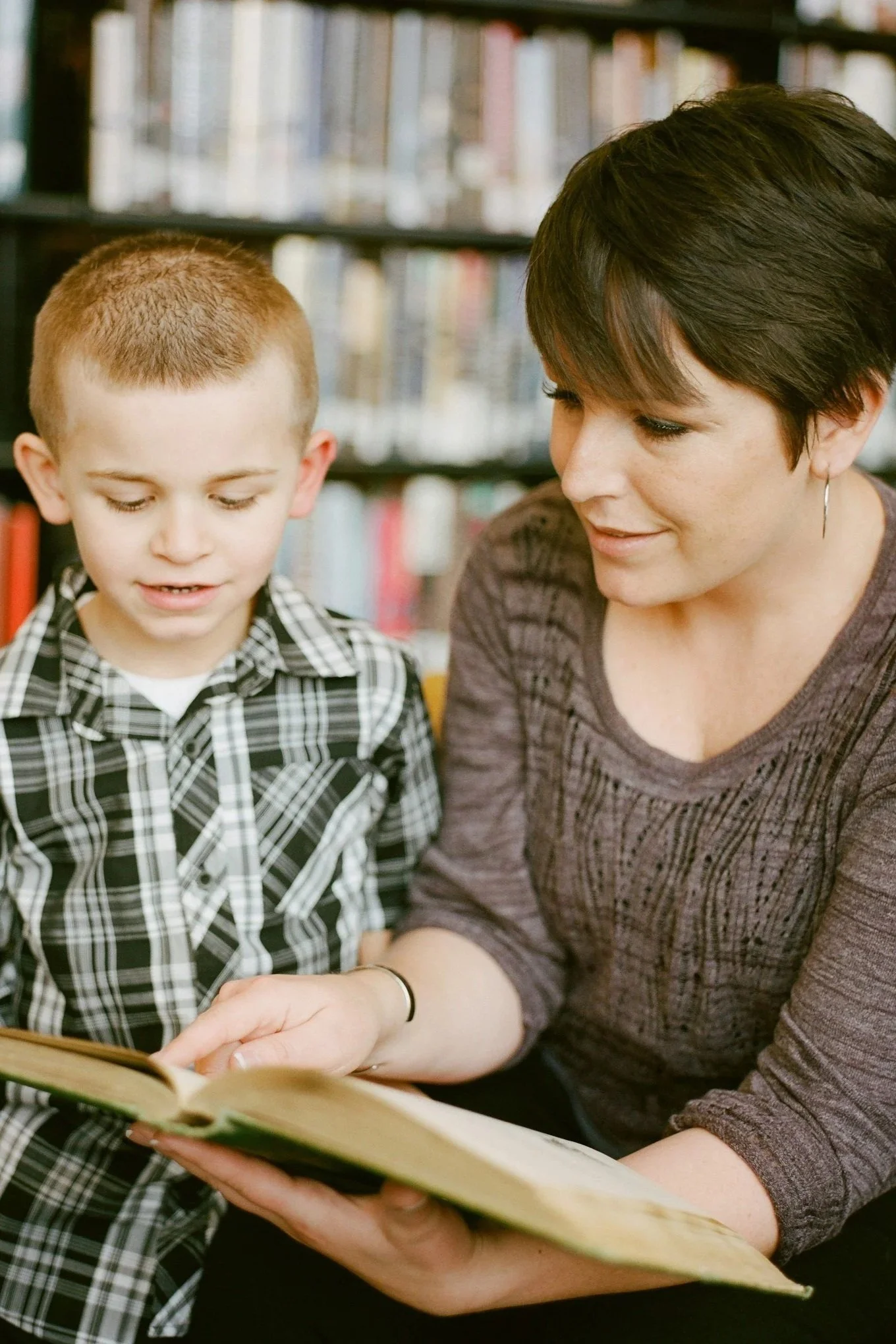 A woman and a young boy sitting together in a library, reading a book. The woman is pointing at the book while the boy looks at it, with bookshelves in the background.