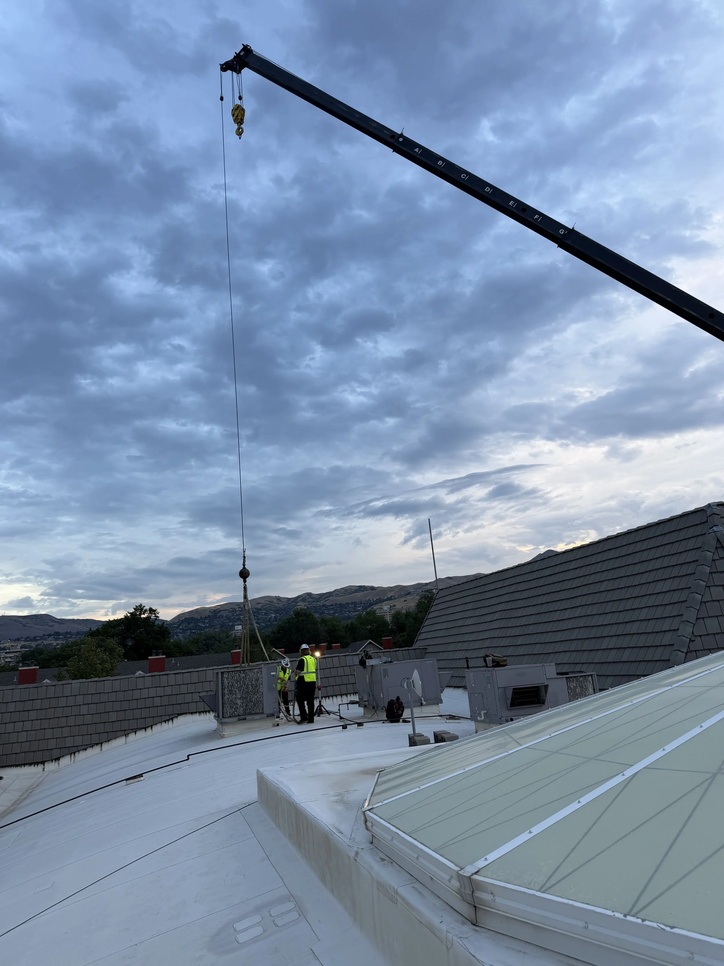 Construction workers on a rooftop near air conditioning units, with a crane in the background and mountains under a cloudy sky.