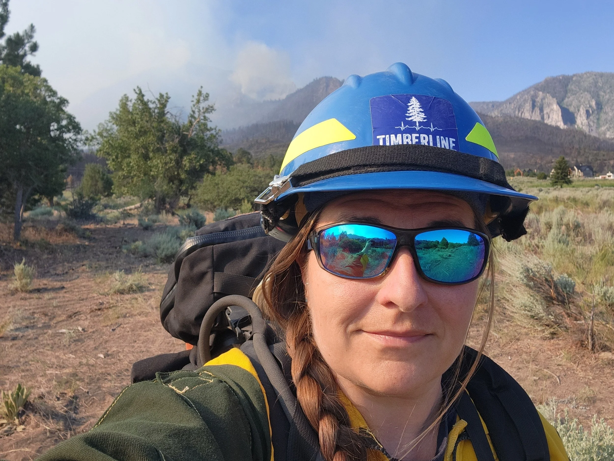 Hiker wearing yellow jacket, sunglasses, and a blue helmet camping in a mountain and forest landscape.