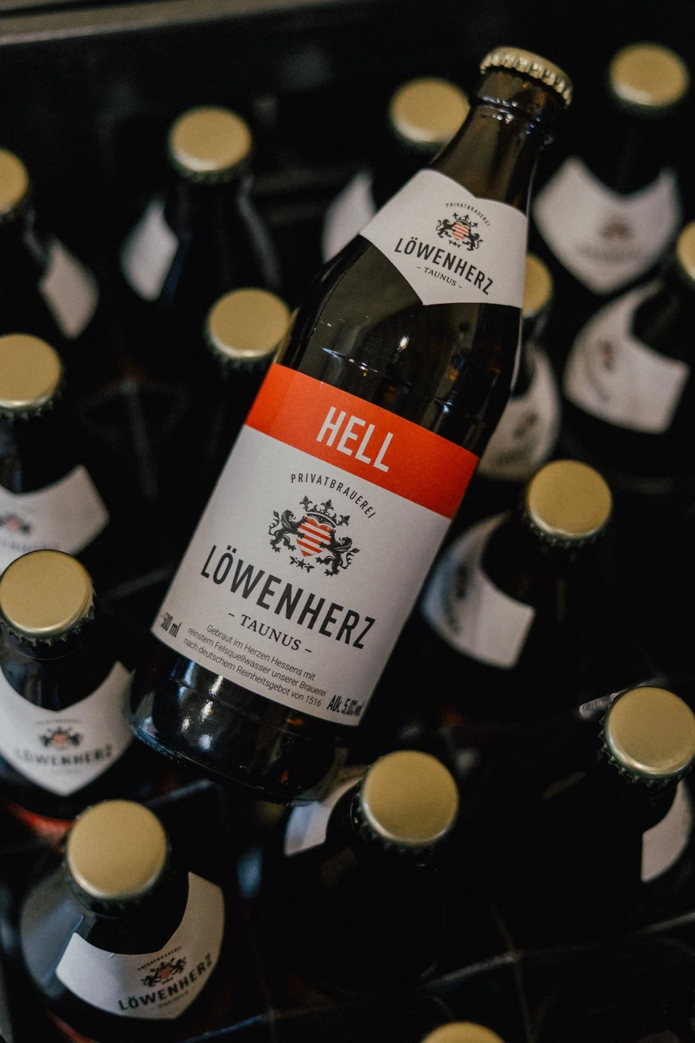 Bierflasche mit dem Etikett 'HELL' von Löwenherz Taunus auf einem Holzgestell mit mehreren weiteren Flaschen im Hintergrund.