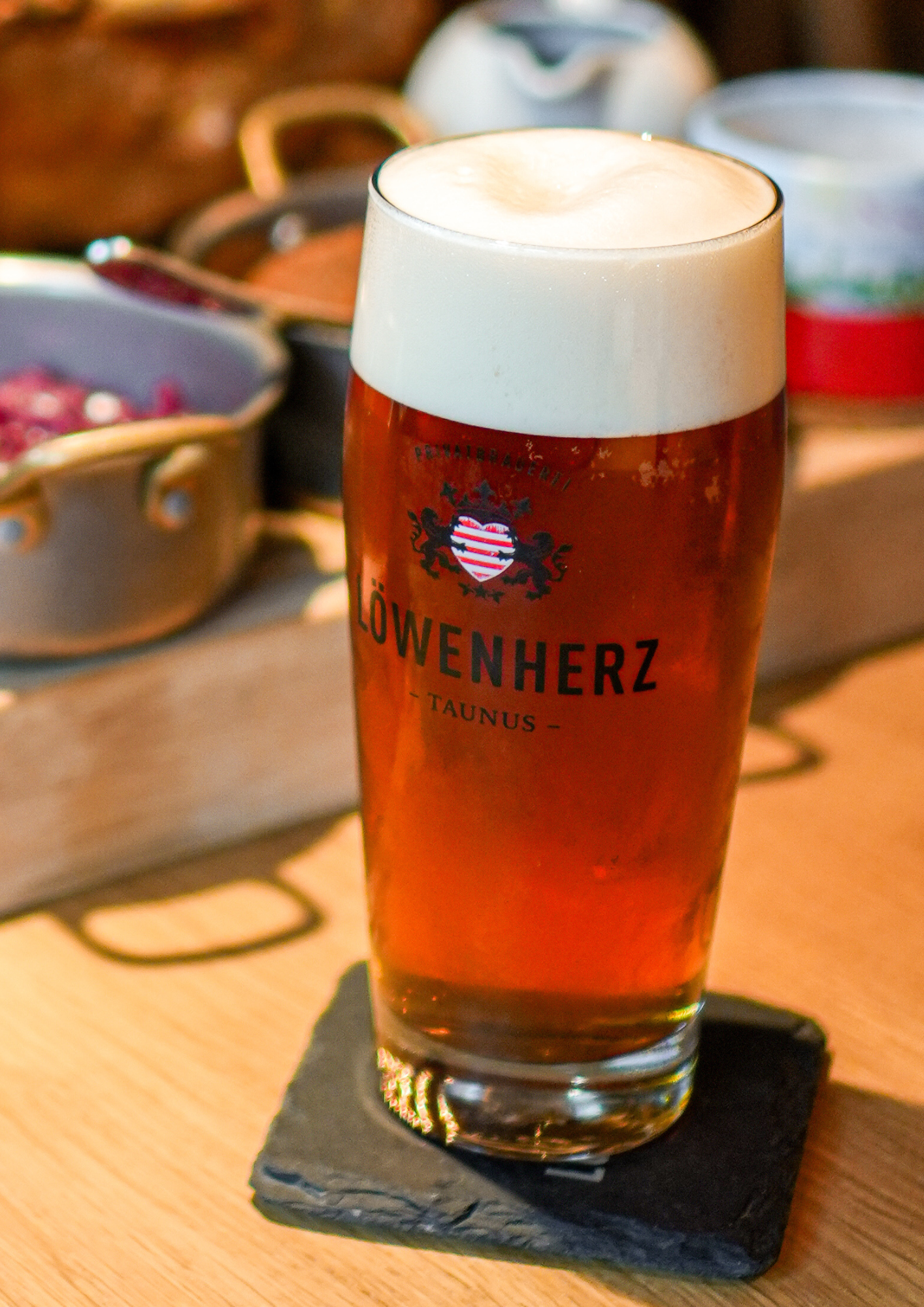 Ein Glas hoppedfenes Bier mit Schaumkrone auf einem schwarzen Untersetzer, im Hintergrund eine Metallbox mit verschiedenen Zutaten. Bier der Löwenherz Privatbrauerei.