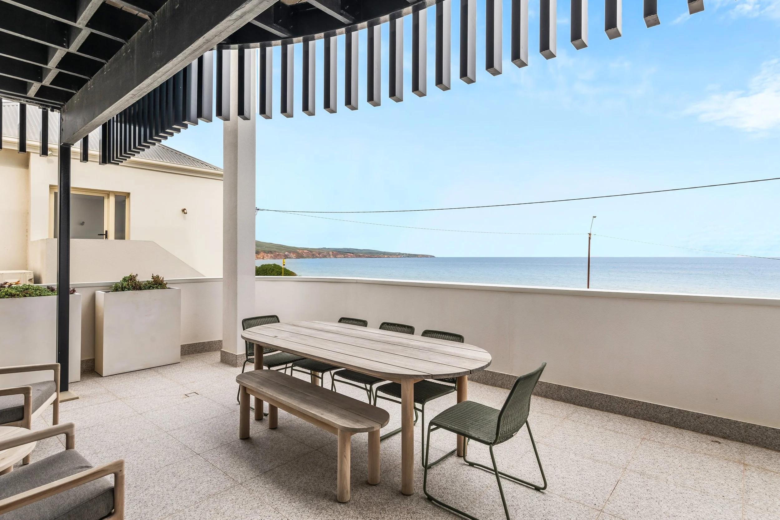 Balcony overlooking the ocean with a wooden table, black chairs, and a white bench, under a black pergola, with a white building on the left and a view of the coastline in the distance under a partly cloudy sky.