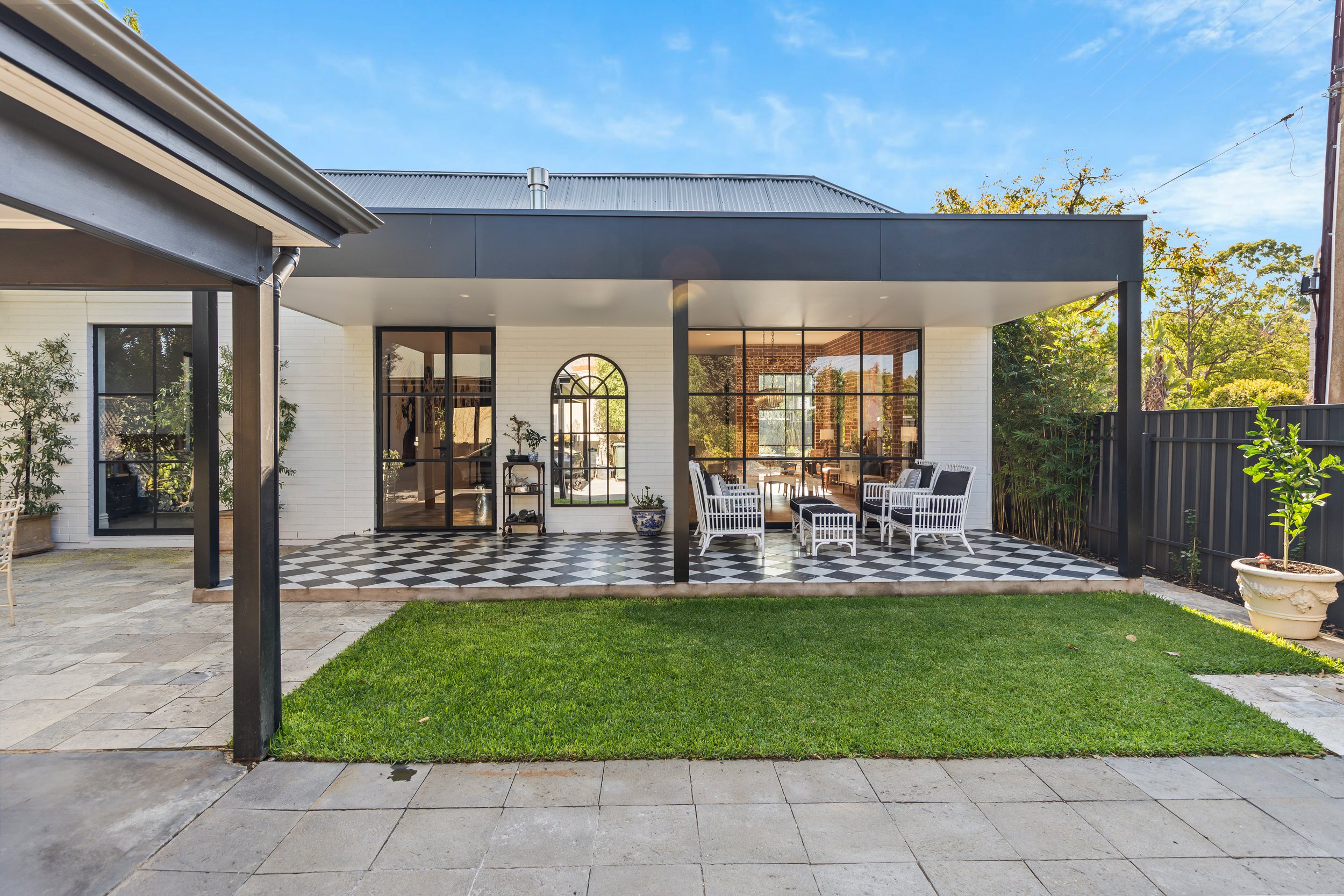 Modern outdoor patio with black and white checkered tiles, white wicker seating, potted plants, and a black fence, attached to a white brick house with large glass windows and sliding doors under a blue sky.