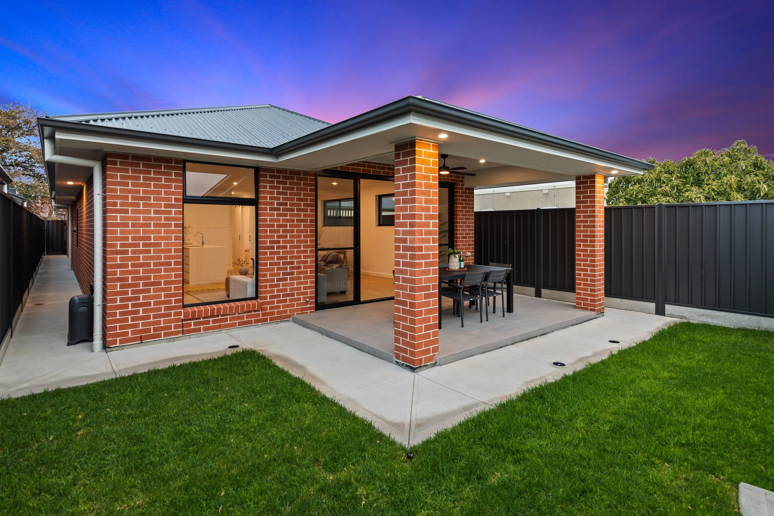 Backyard patio with brick house, sliding glass doors, outdoor dining table, and black privacy fence, during sunset.