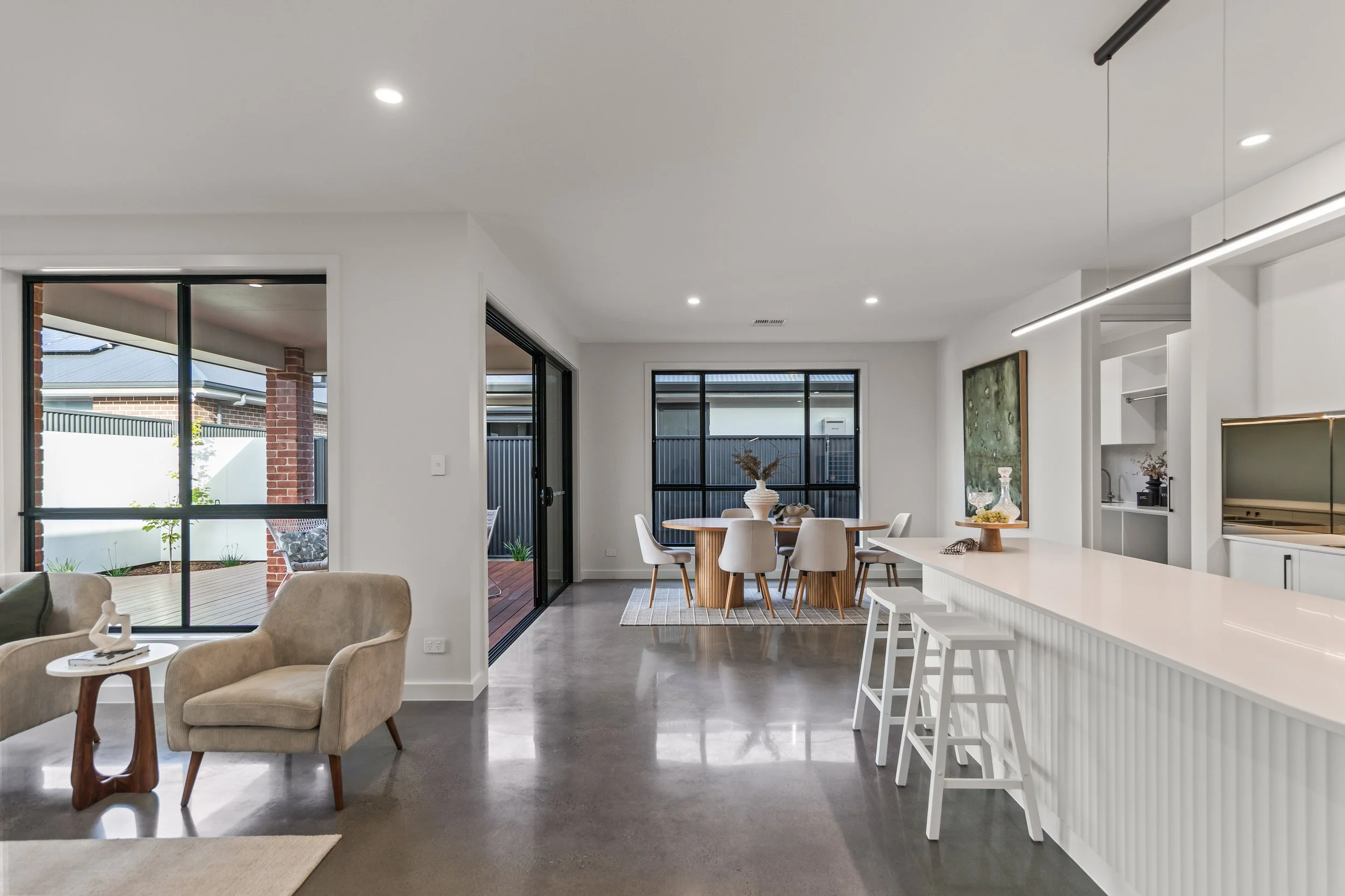 Open-plan living and dining area with white walls, polished concrete floors, large windows, a round dining table with six chairs, a kitchen island with barstools, a beige armchair, and outdoor patio visible through sliding glass doors.