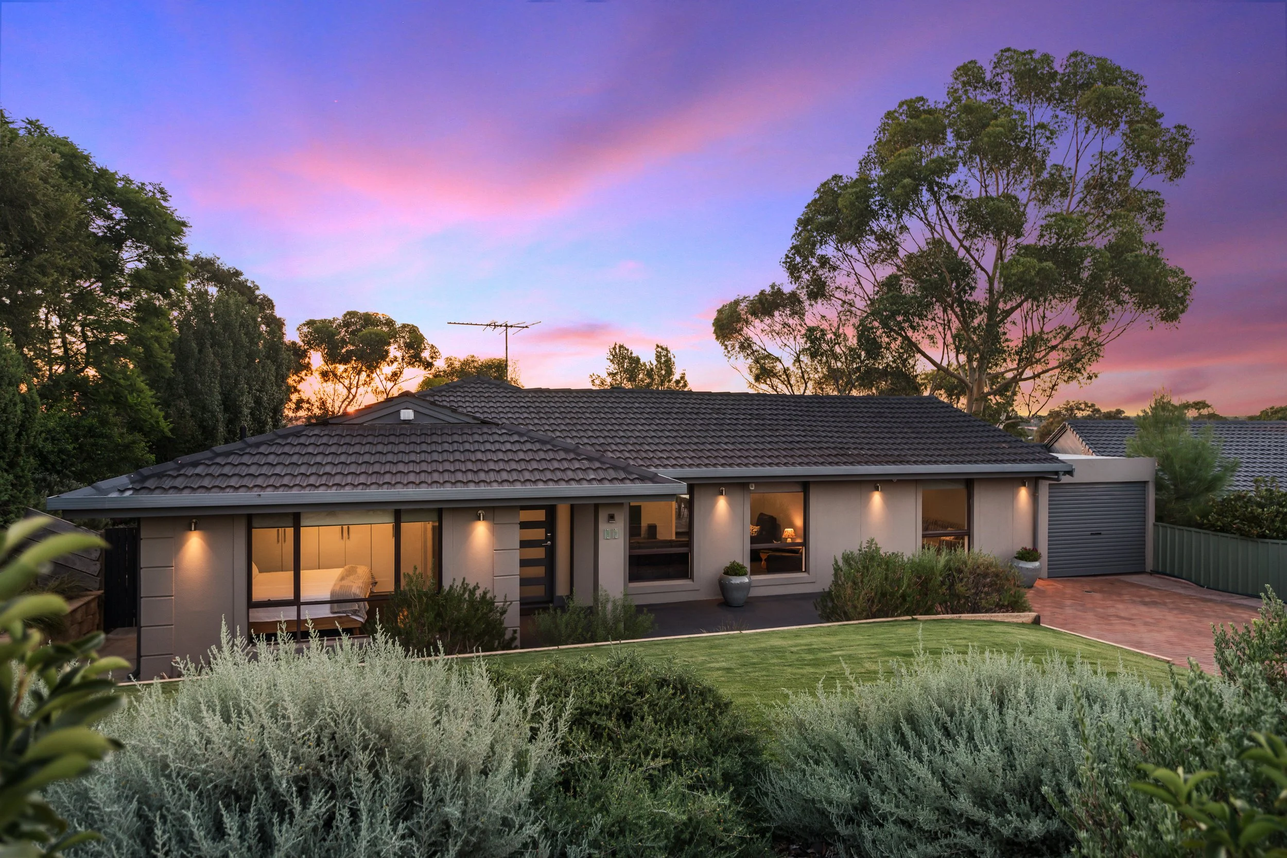 A modern single-story house with a tiled roof and large windows, surrounded by a landscaped garden at dusk with a colorful sky.